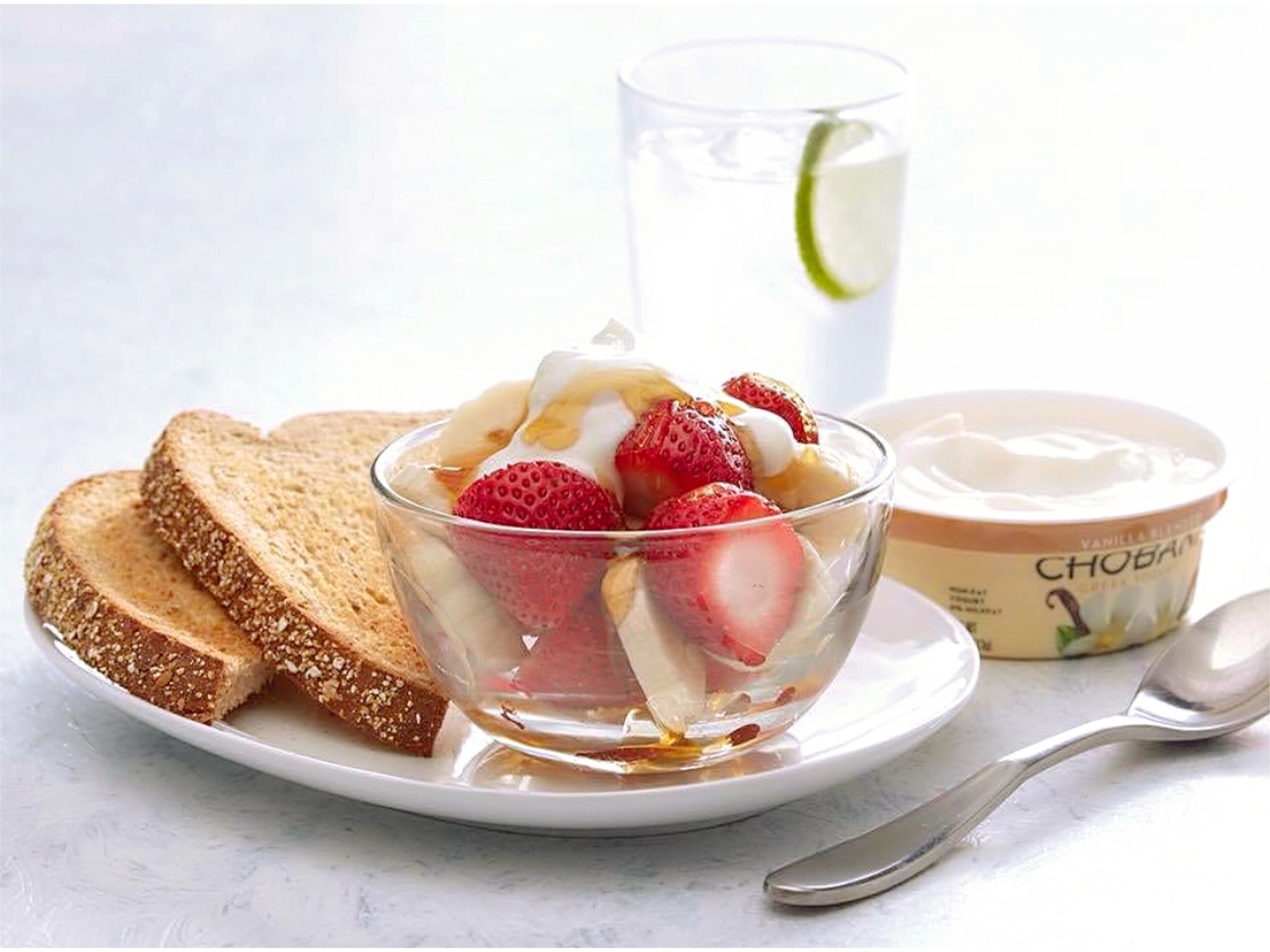A bowl of yogurt with strawberries and banana slices, accompanied by slices of whole grain bread and a glass of water with a lime slice.