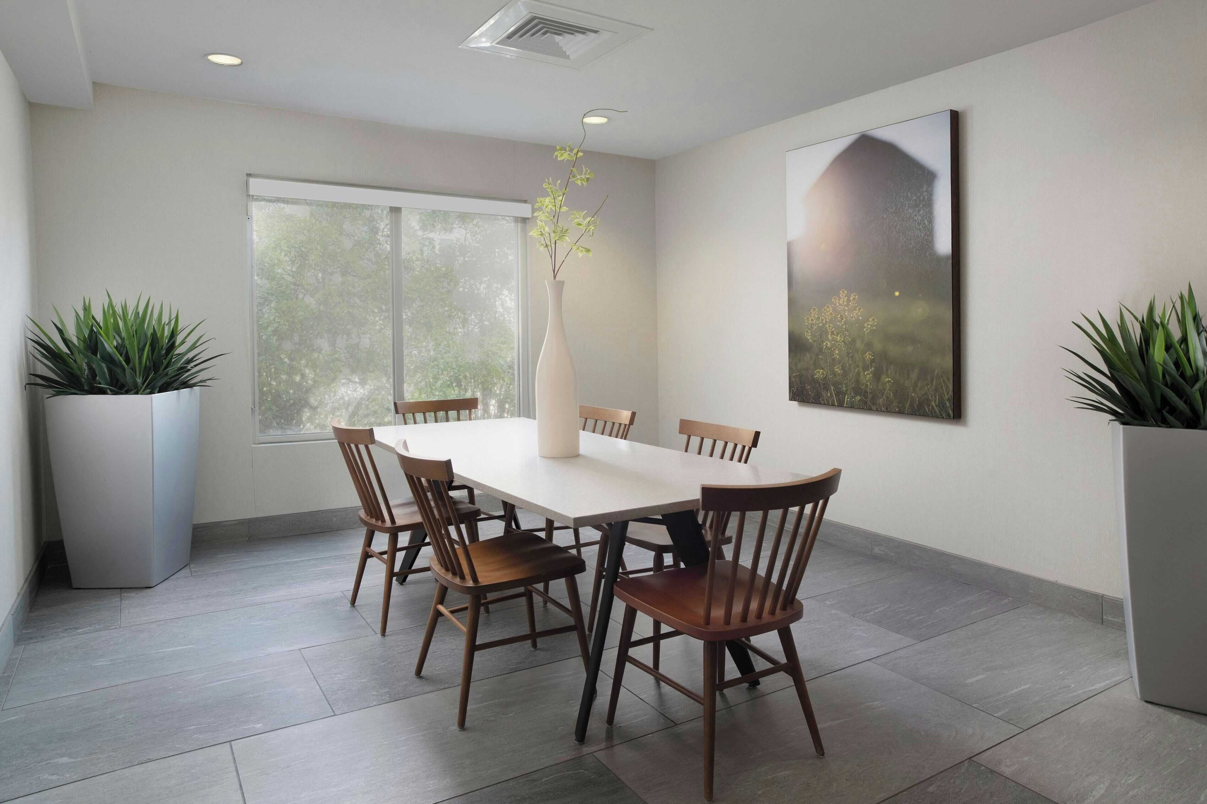 A dining area with a white table, wooden chairs, a vase with flowers, and potted plants.