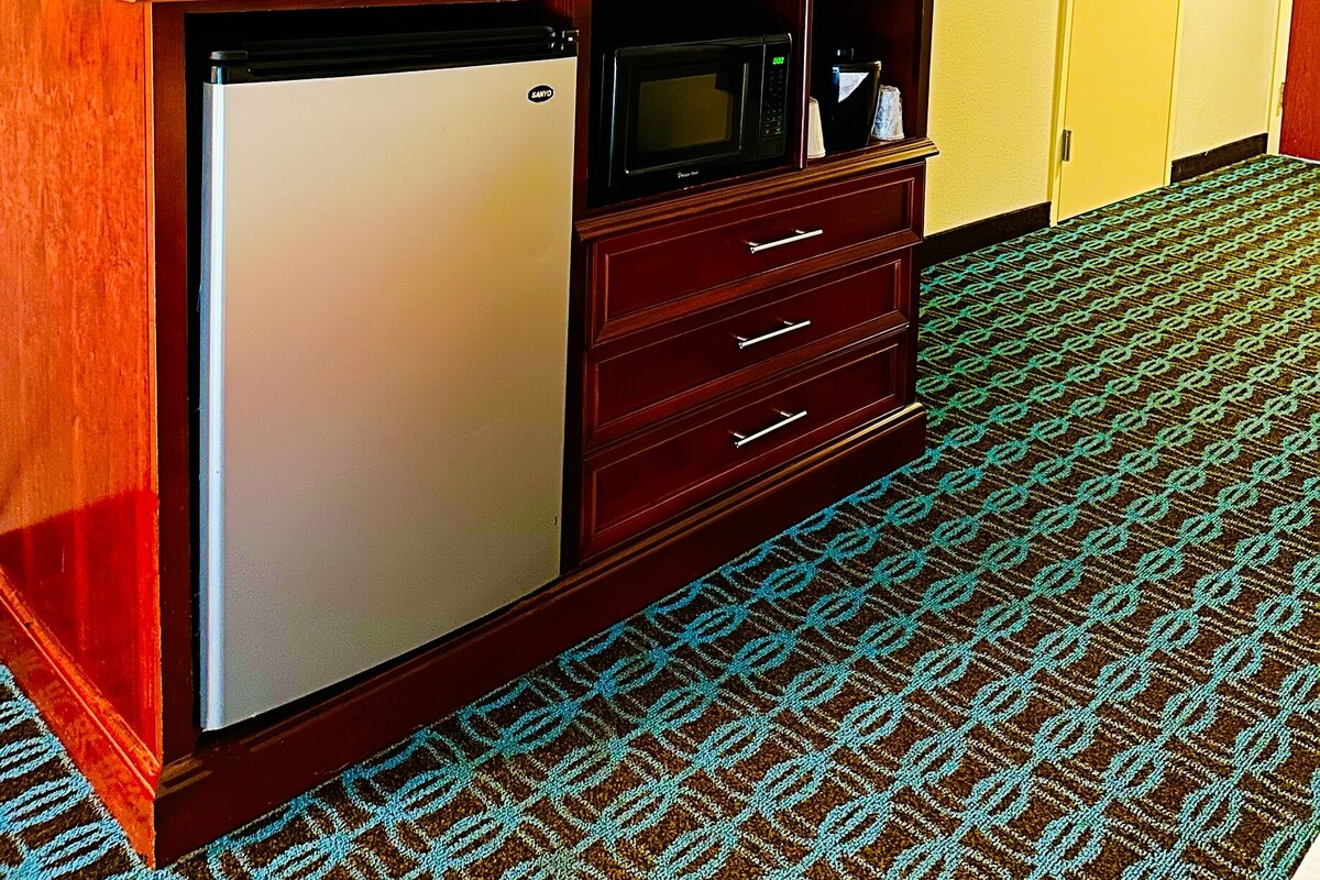 A hotel room with a wooden desk, a television, a mini-fridge, a microwave, and a patterned carpet.
