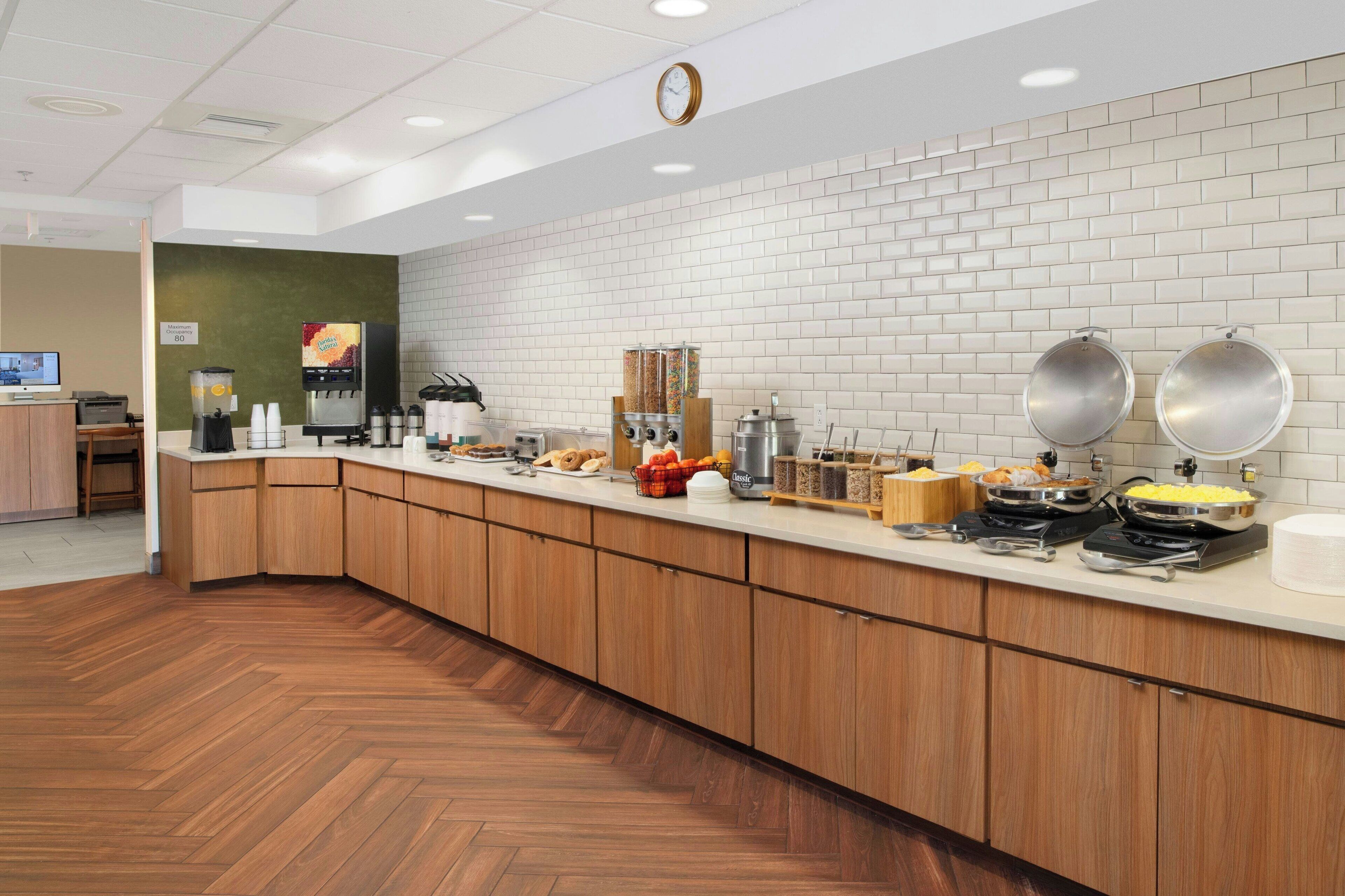 A modern kitchen area with a long counter, various food items, and a clock on the wall.