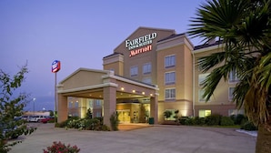 Un hotel Fairfield Inn & Suites Marriott con una entrada bien iluminada, un estacionamiento y una zona ajardinada con flores y una palmera.