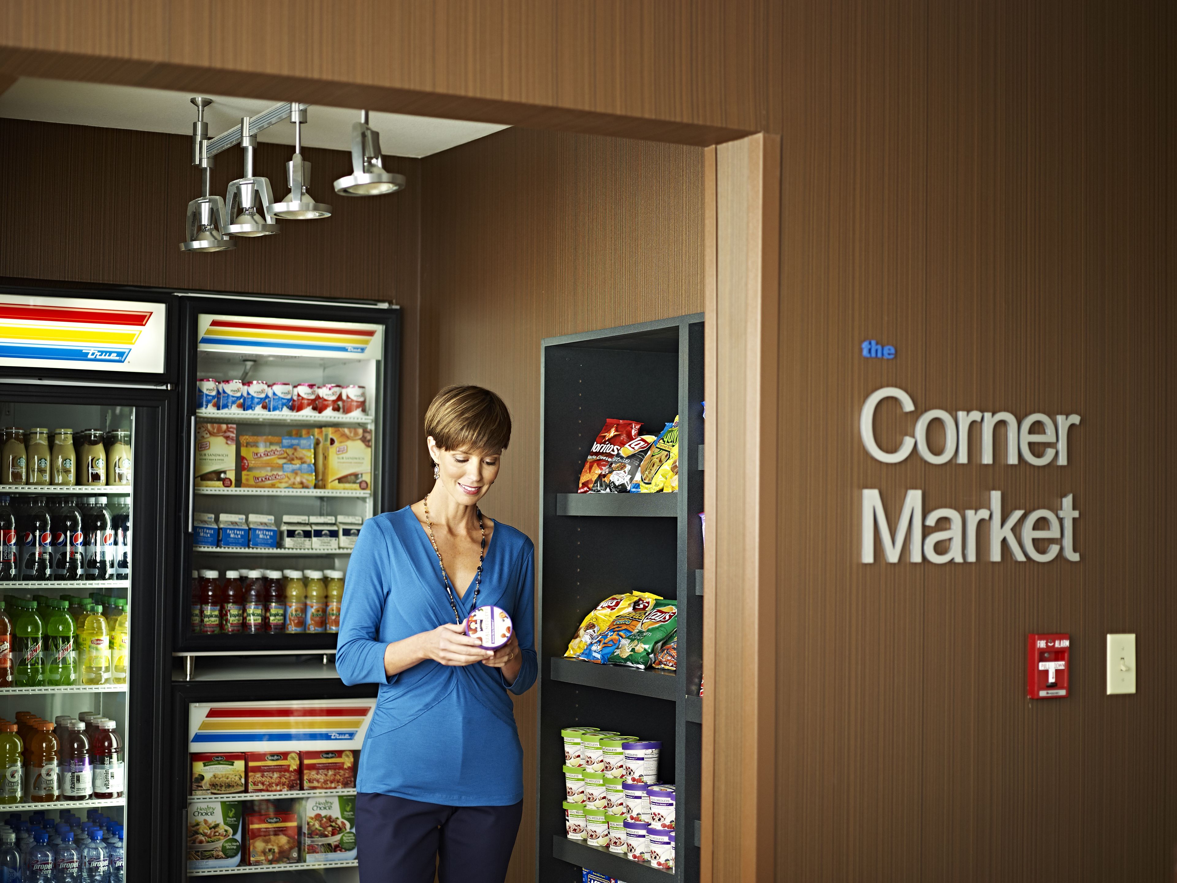 A person in a blue shirt holding a round object, standing in a store with shelves stocked with snacks and beverages, and a refrigerated display case with drinks.