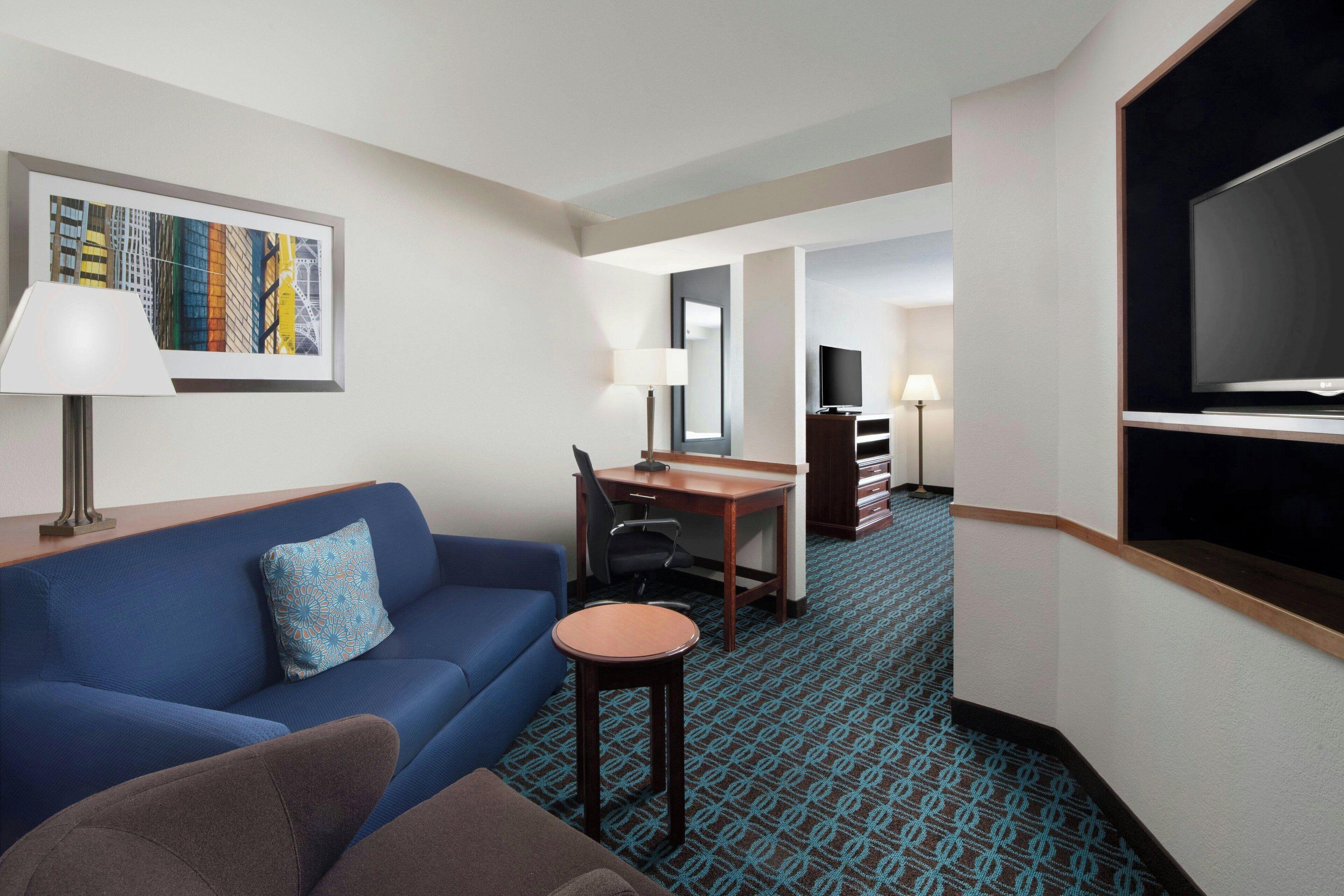 A hotel room with a blue sofa, a small round table, a desk with a chair, a television, and a framed picture on the wall.