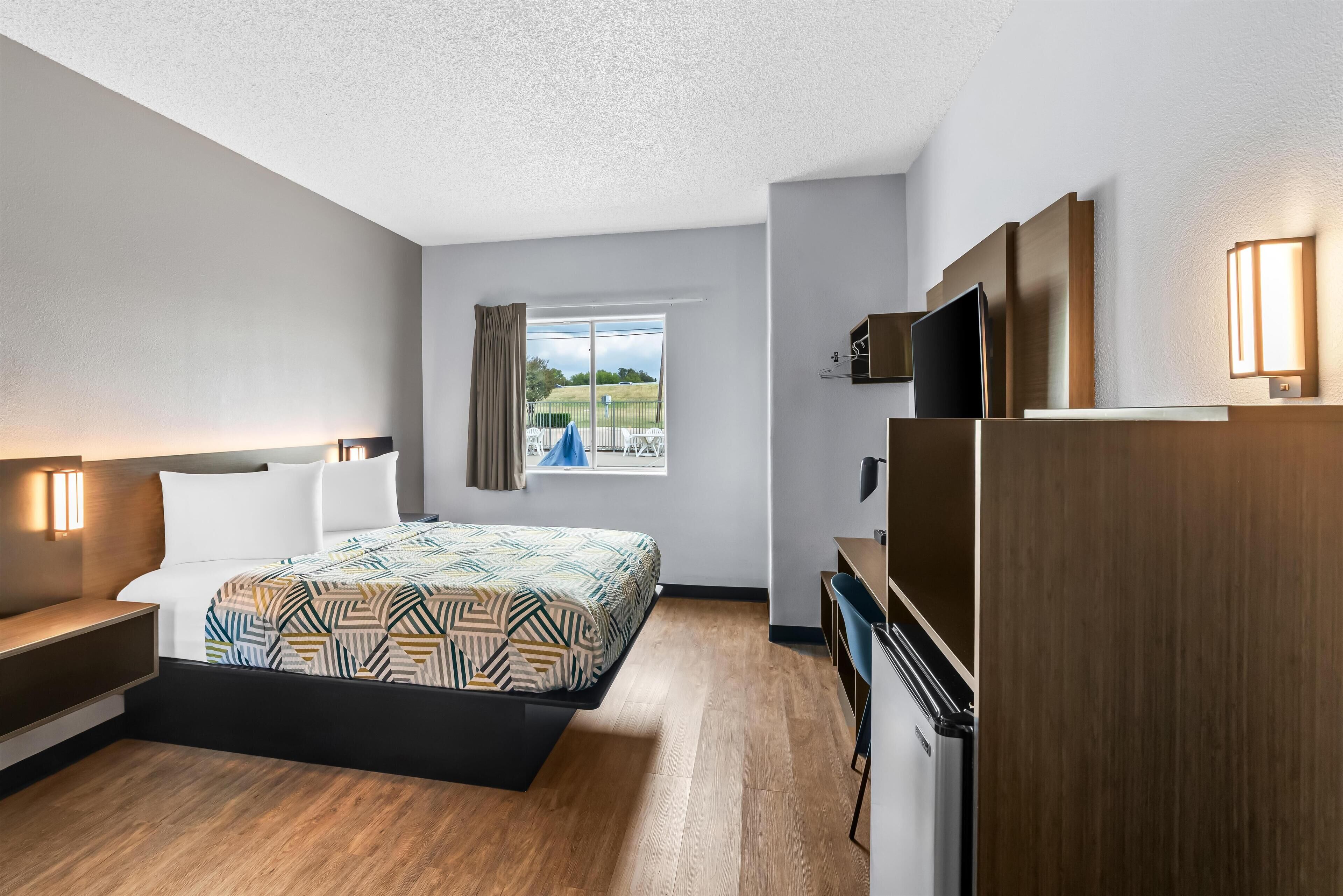 Ein modernes Hotelzimmer mit Bett, Nachttisch, Fernseher und Blick ins Freie.