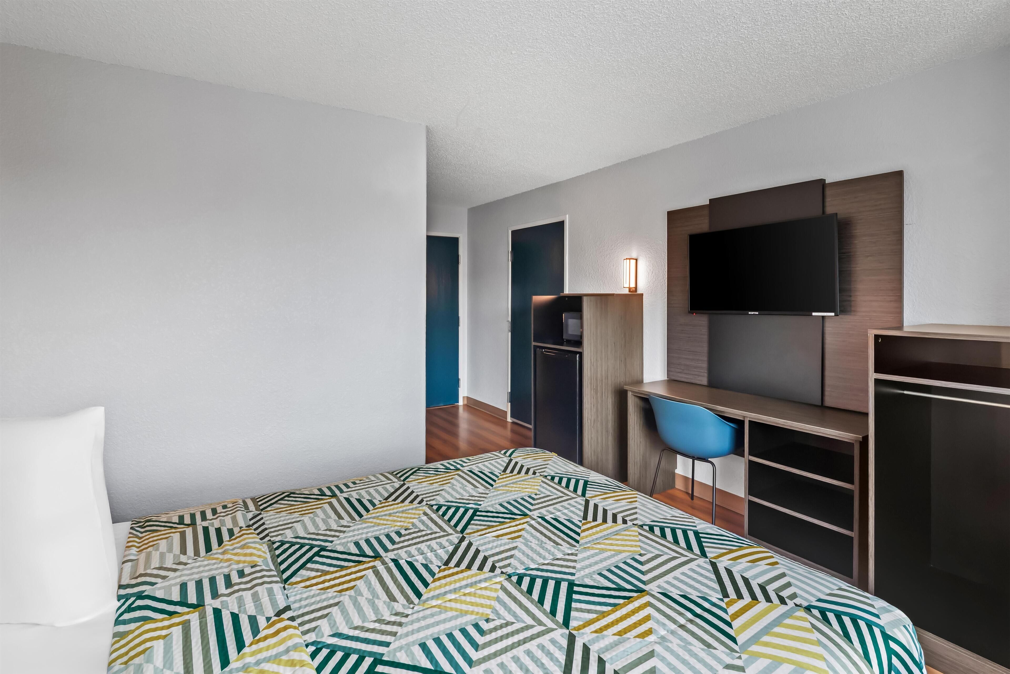 A modern hotel room with a bed, a desk, a chair, a TV, and a wardrobe.