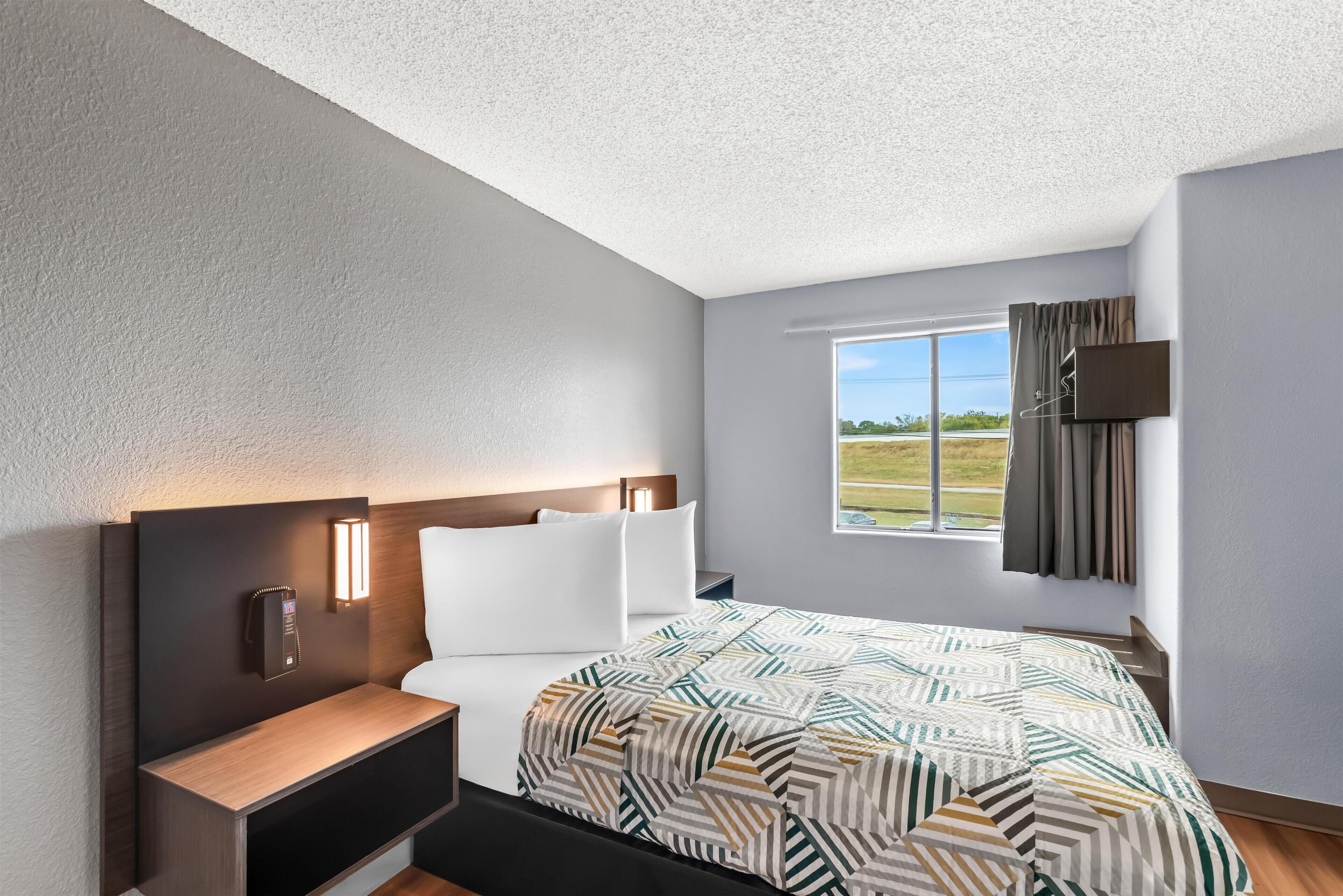 A modern hotel room with a large bed, a bedside table, a nightstand, a wall-mounted TV, and a window with a view of the outdoors.
