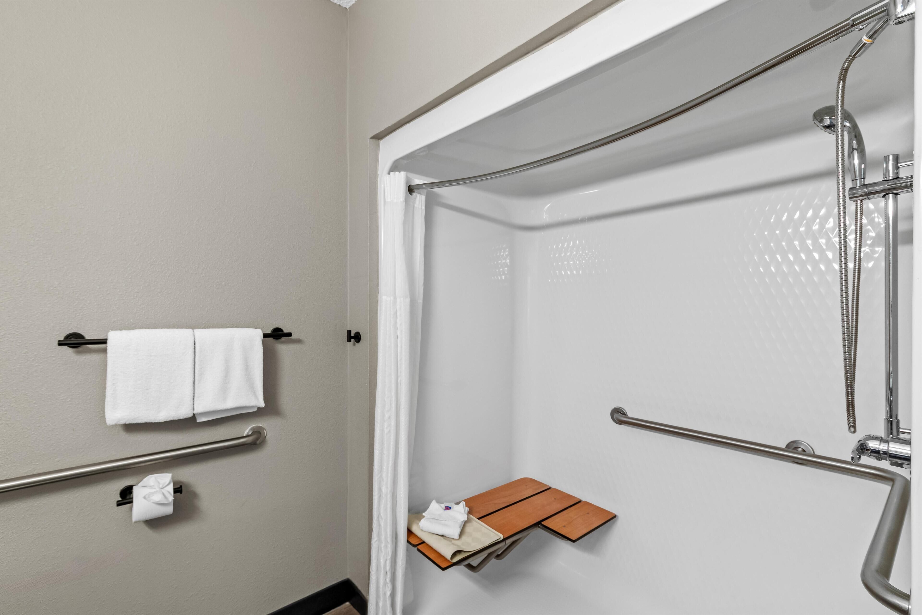 A modern bathroom with a shower, grab bars, a towel rack, and a small wooden shelf.