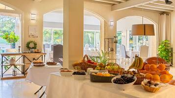 A breakfast buffet with various fruits and pastries, a white tablecloth, and a view of the outdoors through large windows.