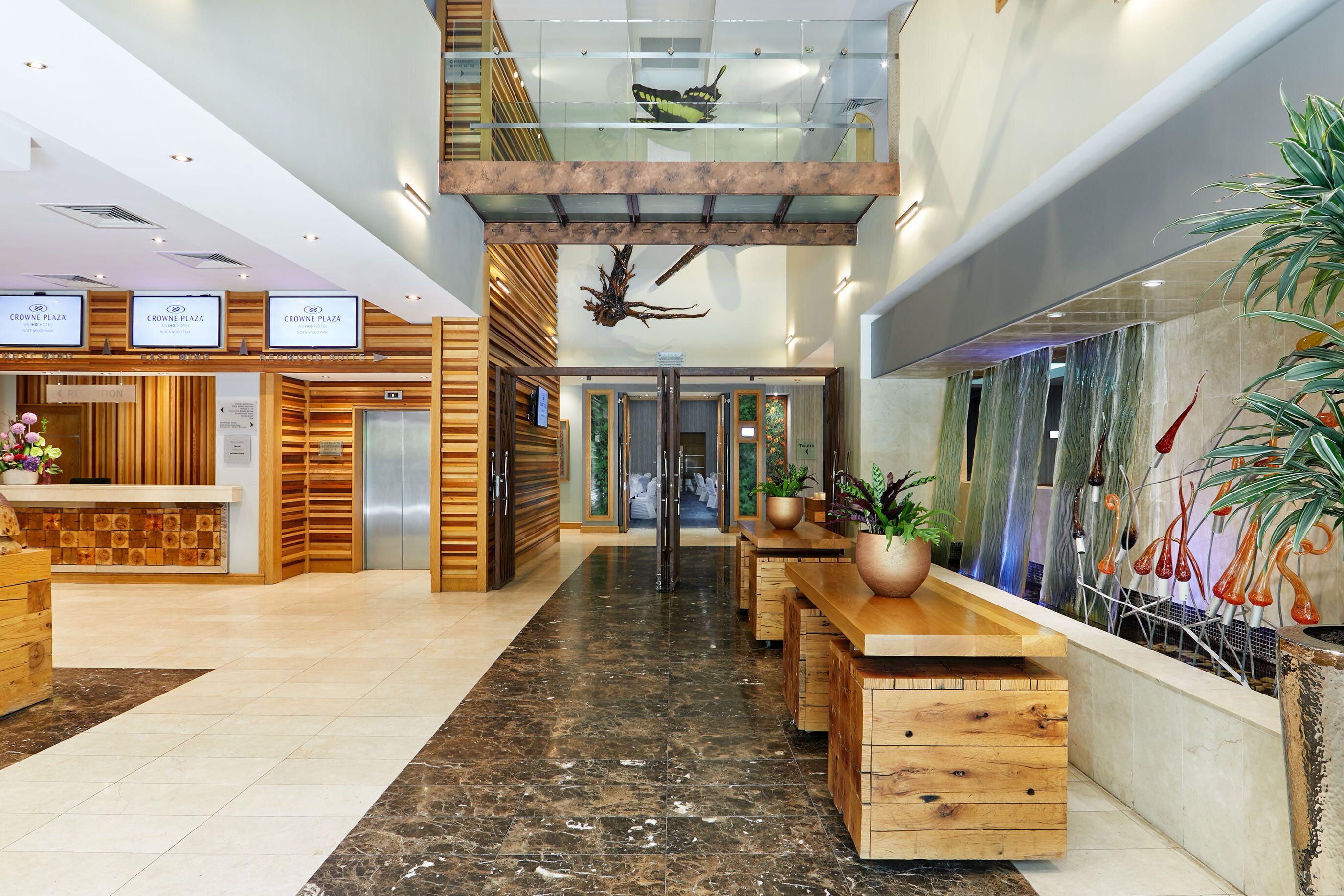 A hotel reception area with a modern design, featuring a reception desk, a staircase, and a seating area with wooden tables and chairs.
