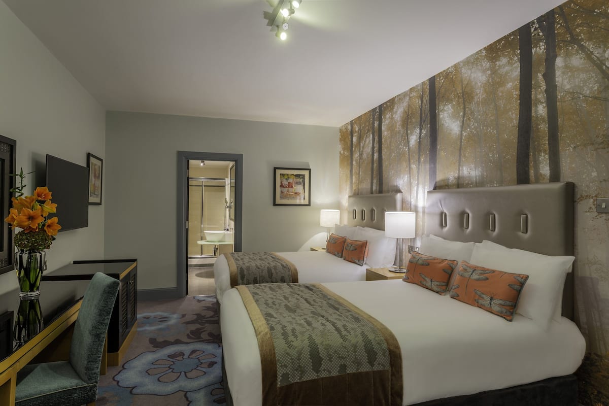 A hotel room with two beds, a desk, a chair, a vase with flowers, and a painting on the wall.