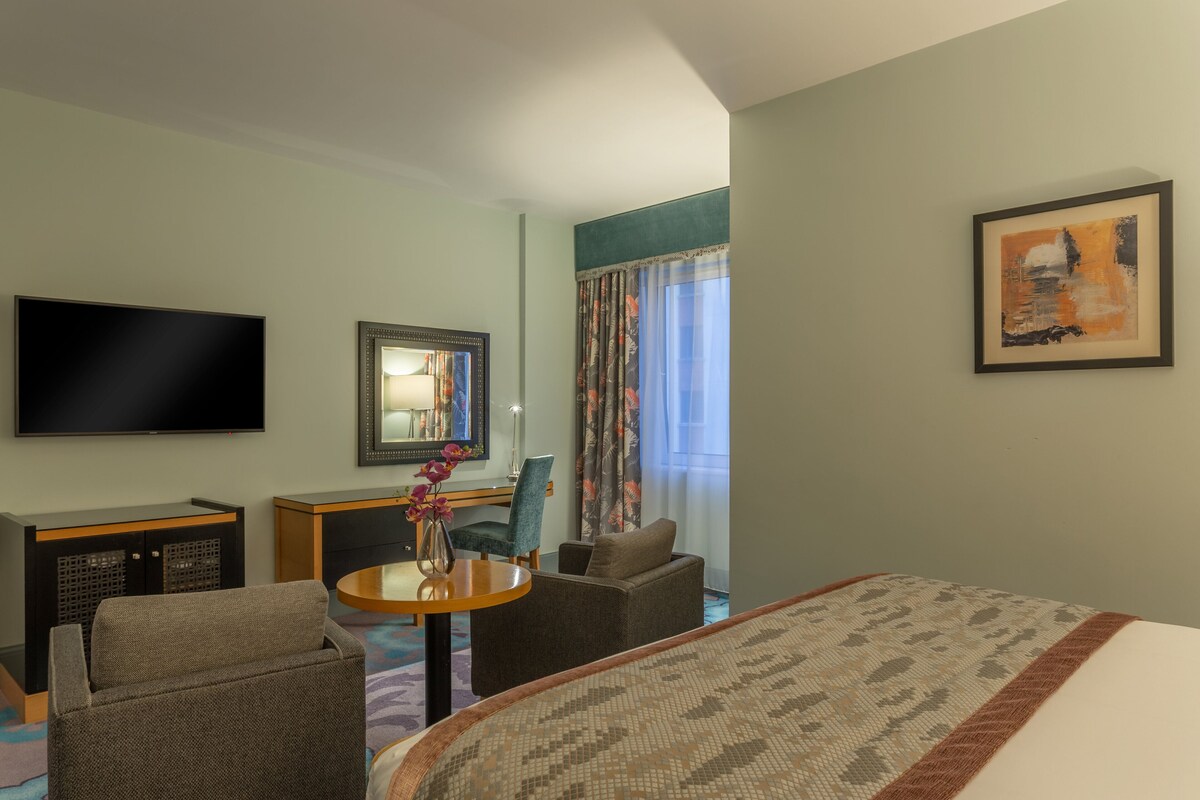Une chambre d’hôtel comprenant un lit, une télévision, un bureau avec une chaise, une petite table avec un vase et une fenêtre avec des rideaux.