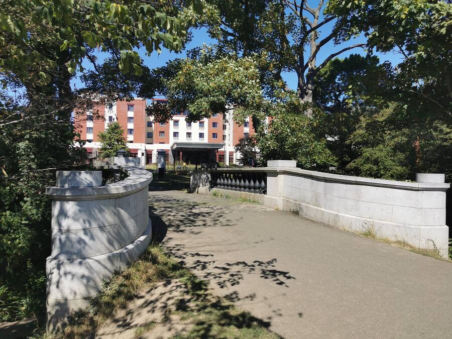 A paved pathway with a stone wall on one side and a building with multiple windows in the background.