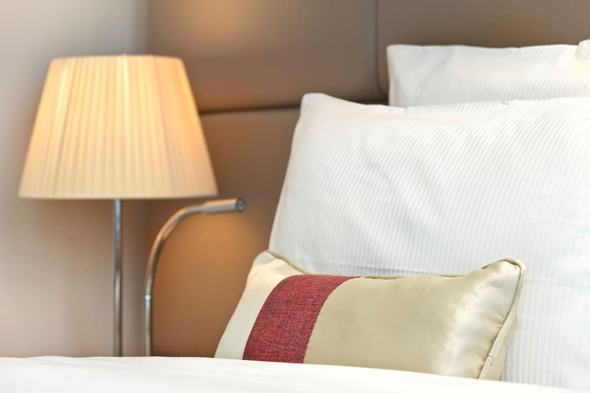 A neatly made bed with white linens and a red accent pillow, a bedside lamp, and a headboard.
