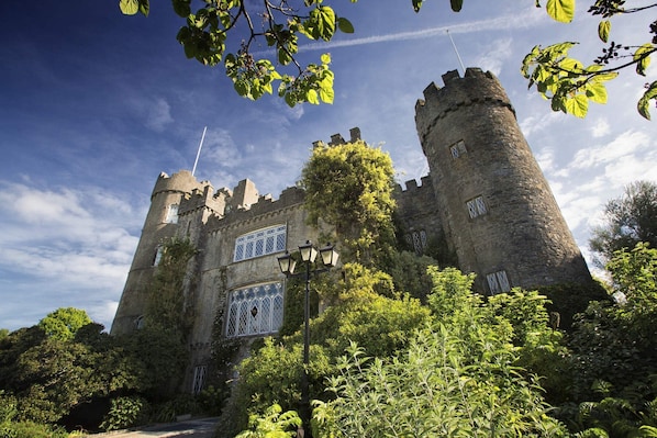A historic castle with two prominent towers, a large arched window, and lush greenery surrounding the structure.