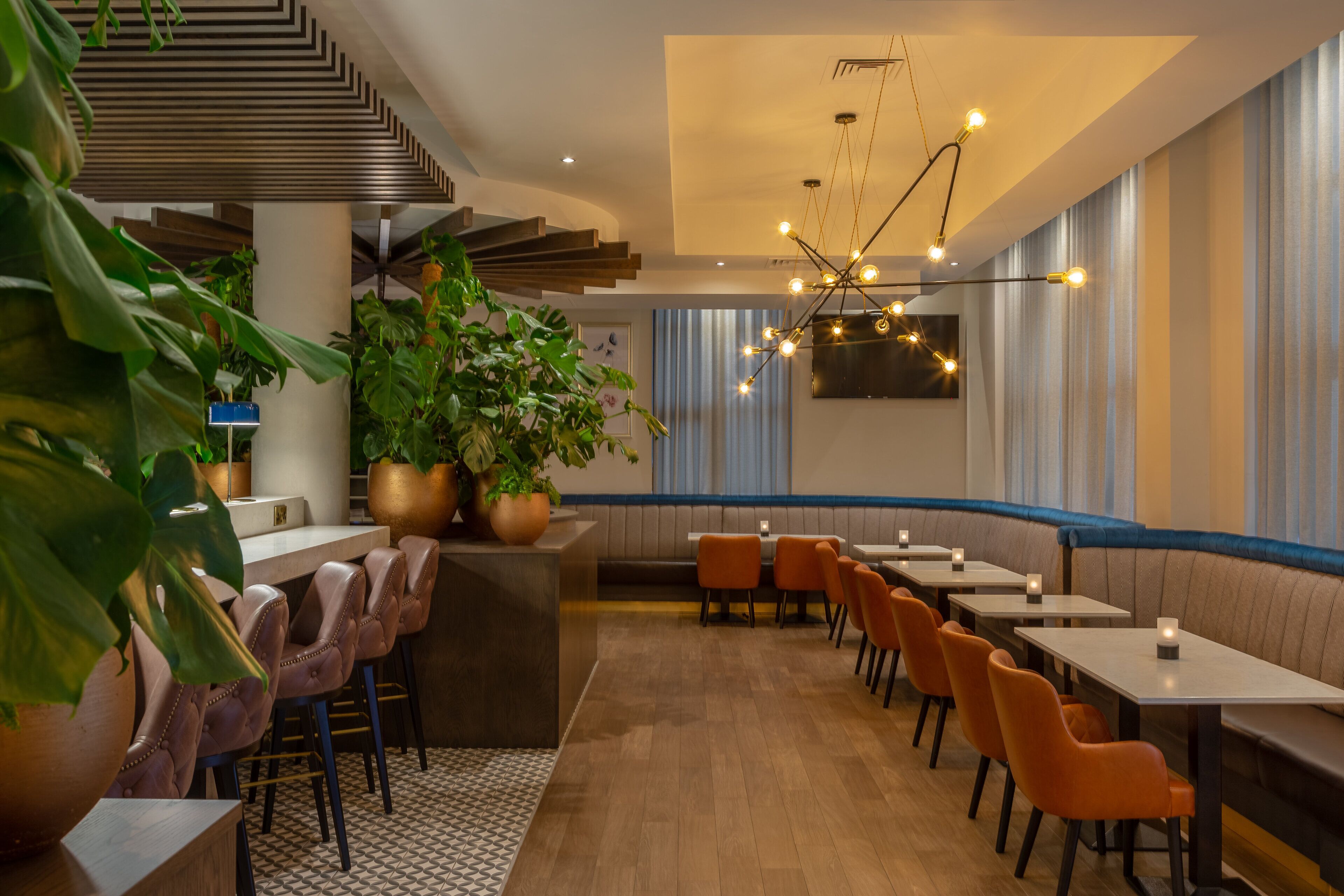 A modern restaurant with a central bar, seating along the walls, and a staircase leading to an upper level.