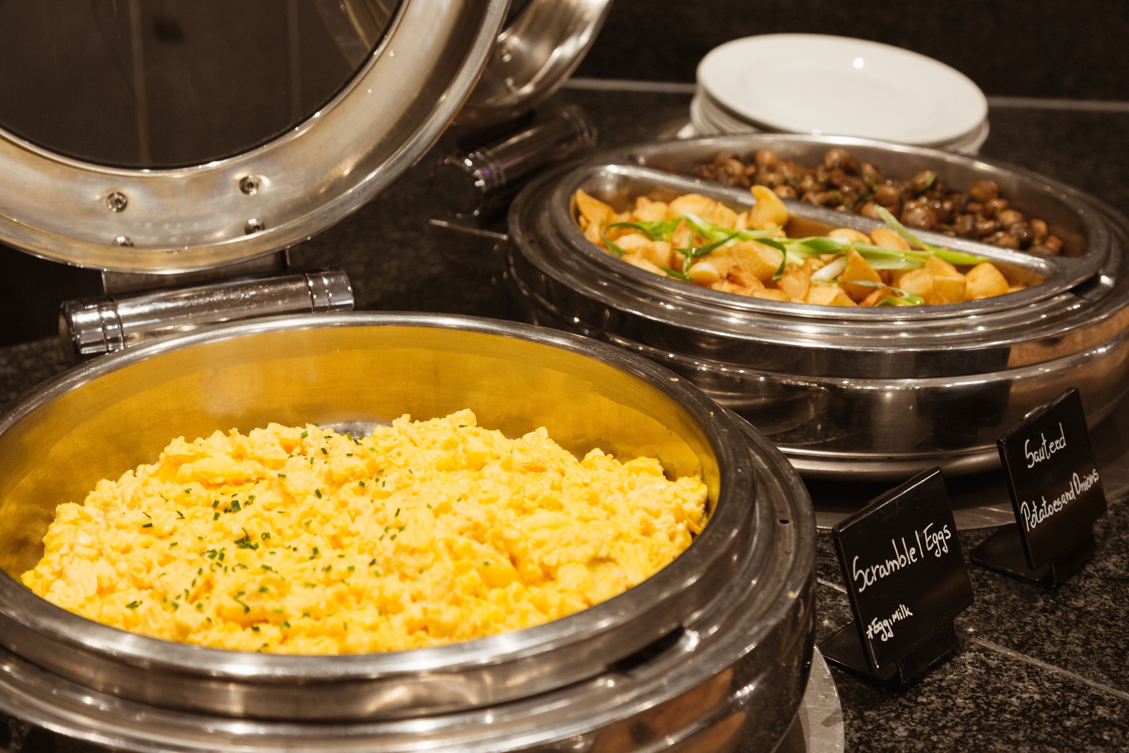 A buffet with labeled dishes including scrambled eggs, sausage, and potatoes.