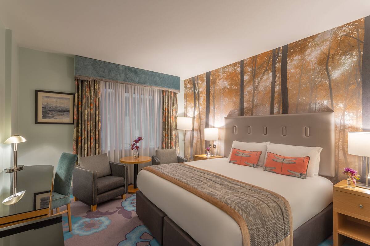 A hotel room with a large bed, two bedside tables, a desk with a chair, a small round table with a chair, and a large window with curtains.