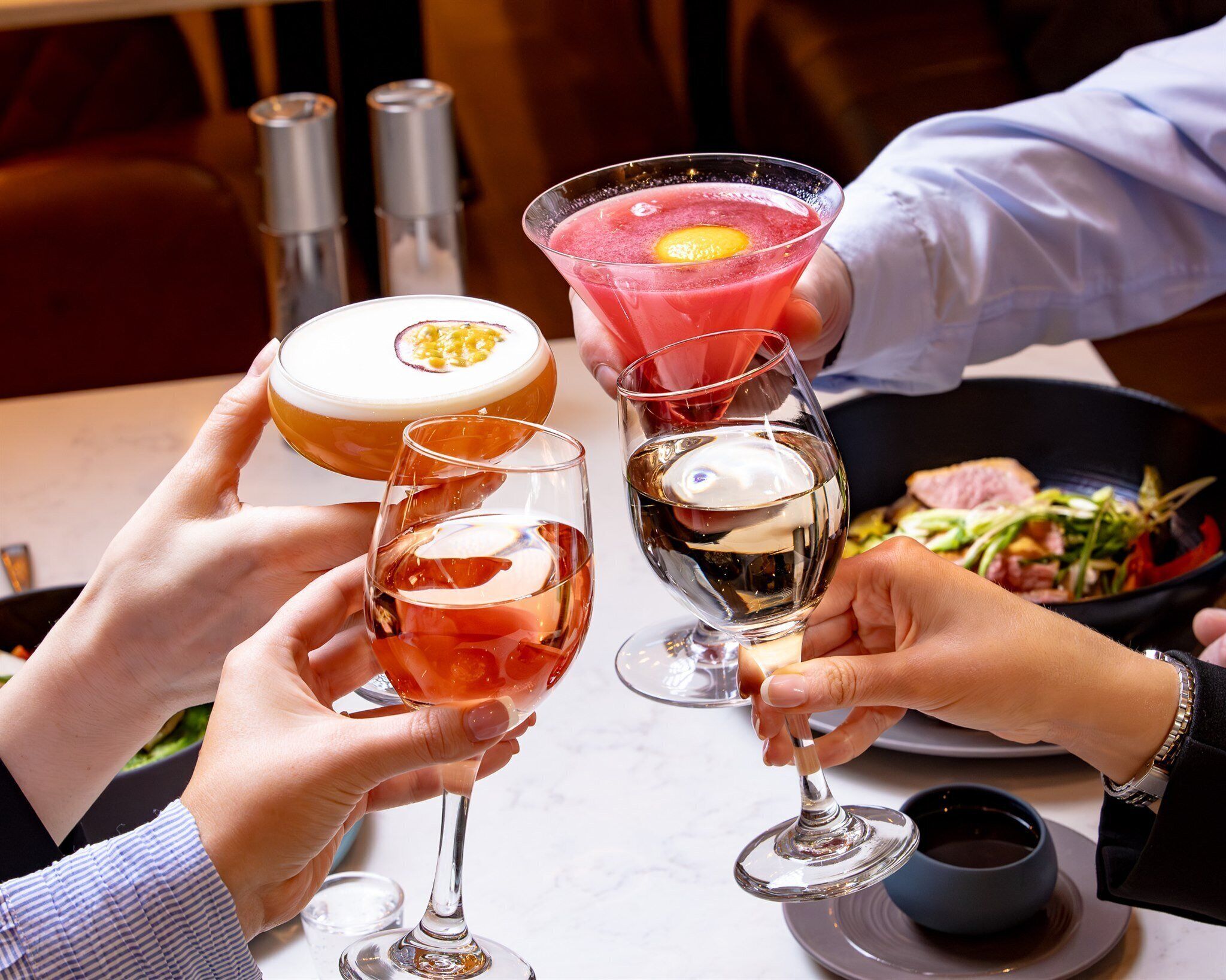A group of people toasting with various drinks, including a cocktail with a fruit garnish, a glass with a creamy beverage, and a wine glass.