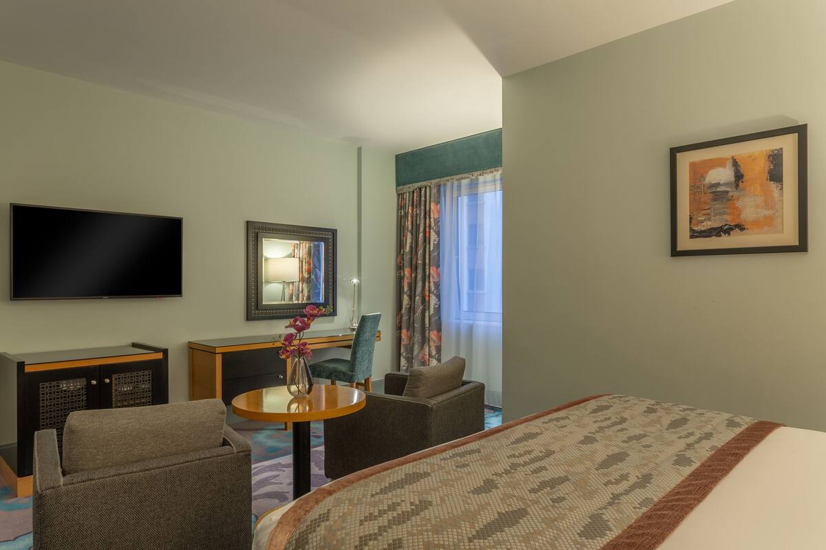 Habitación de hotel con cama, televisión, escritorio con silla, una mesita con un jarrón y ventana con cortinas.
