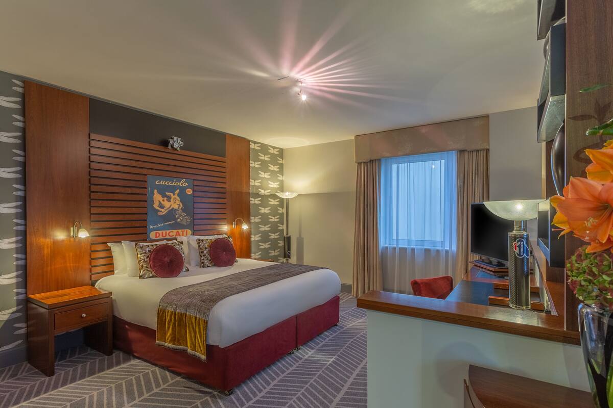 A hotel room with a large bed, a wooden headboard, a nightstand, a desk, and a large window with curtains.