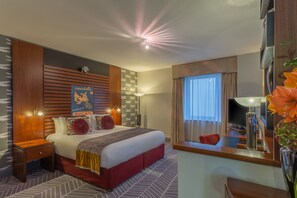 A hotel room with a large bed, a wooden headboard, a nightstand, a desk, and a large window with curtains.