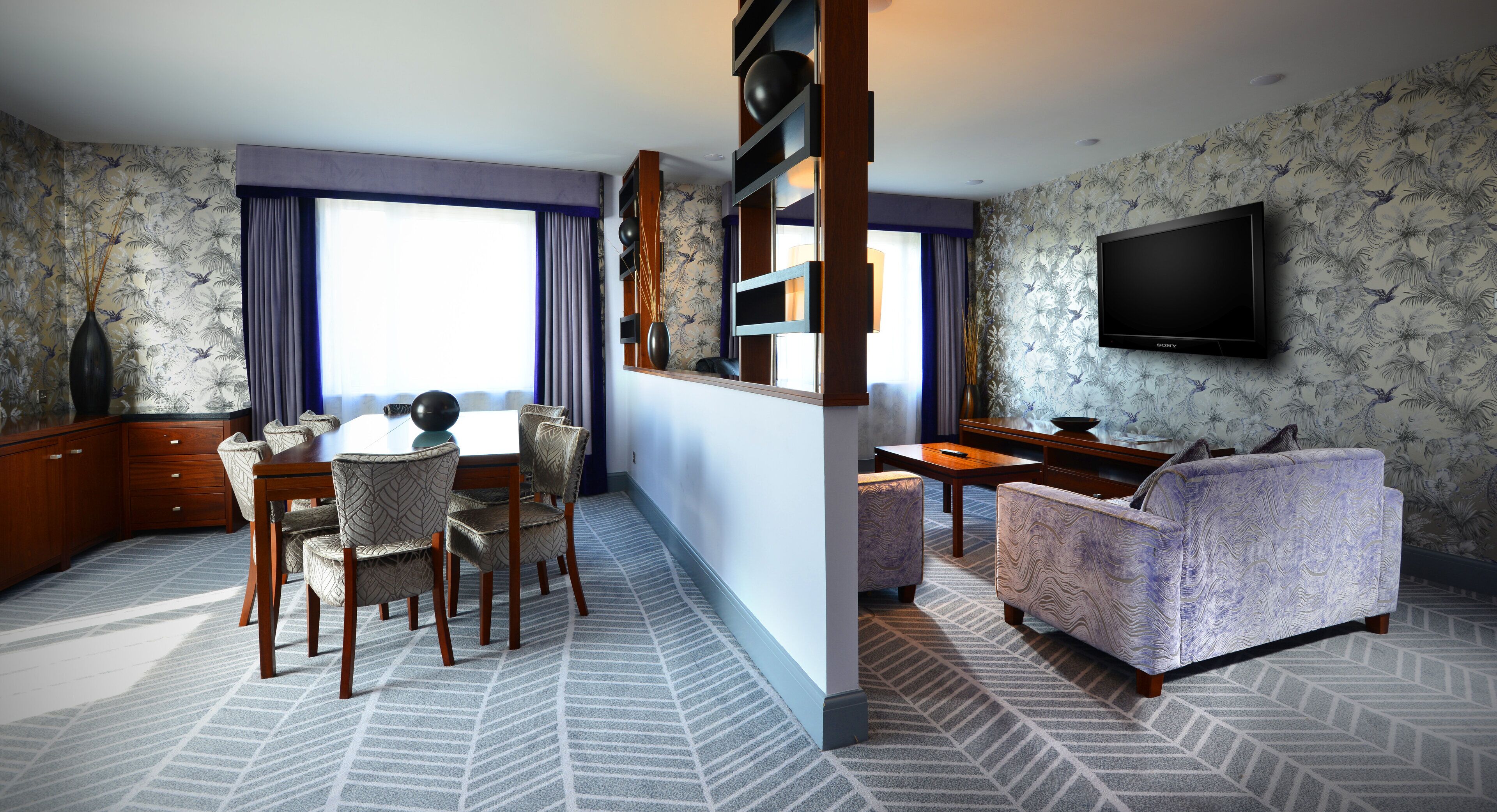 A hotel room with a dining area, a sofa, a TV, and a patterned carpet.