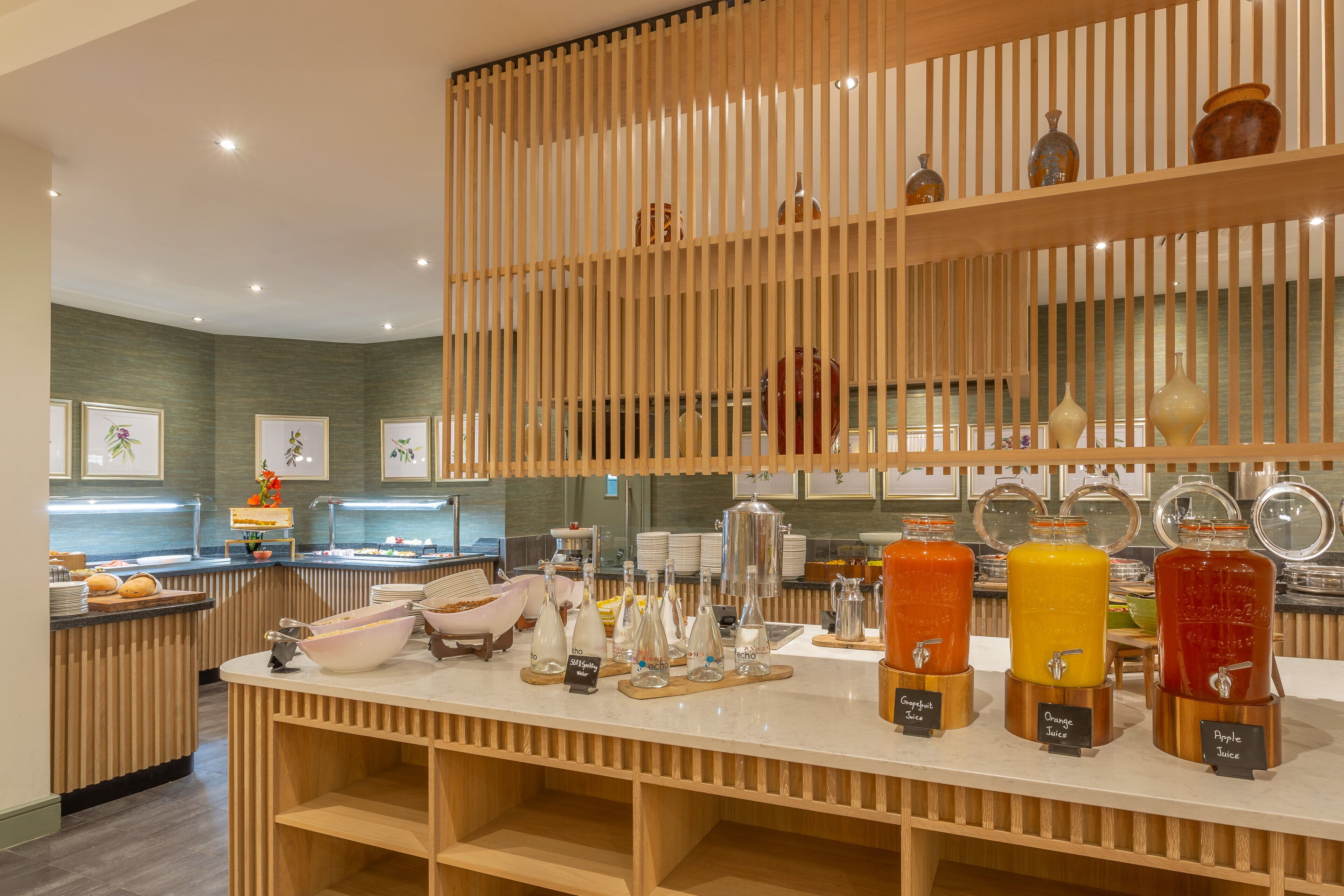 Una barra de desayuno con varios dispensadores de jugo, un panel divisor de madera con listones y un mostrador con alimentos.