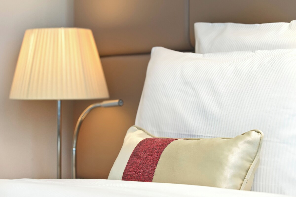 Un lit bien fait, avec du linge de lit blanc et un coussin rouge, une lampe de chevet et une tête de lit.