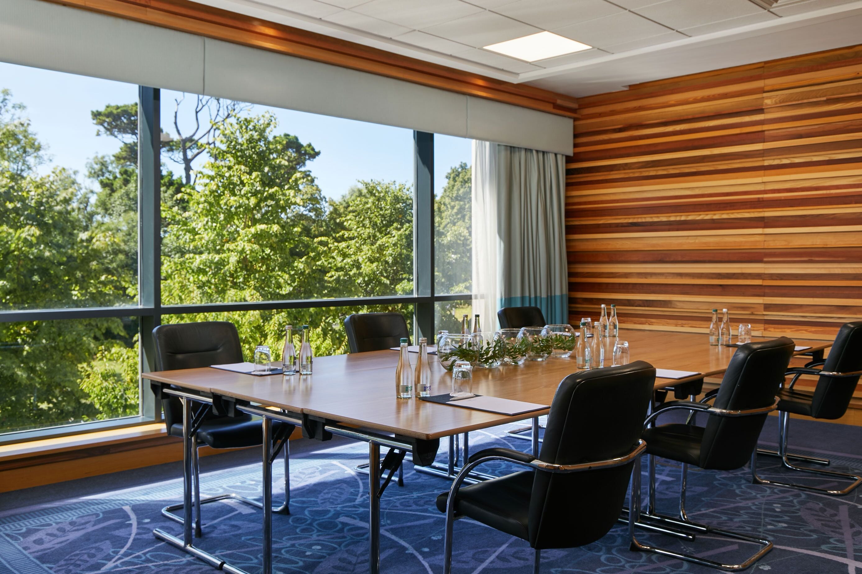 Un salón de conferencias con una gran mesa, sillas y vistas a árboles a través de amplias ventanas.