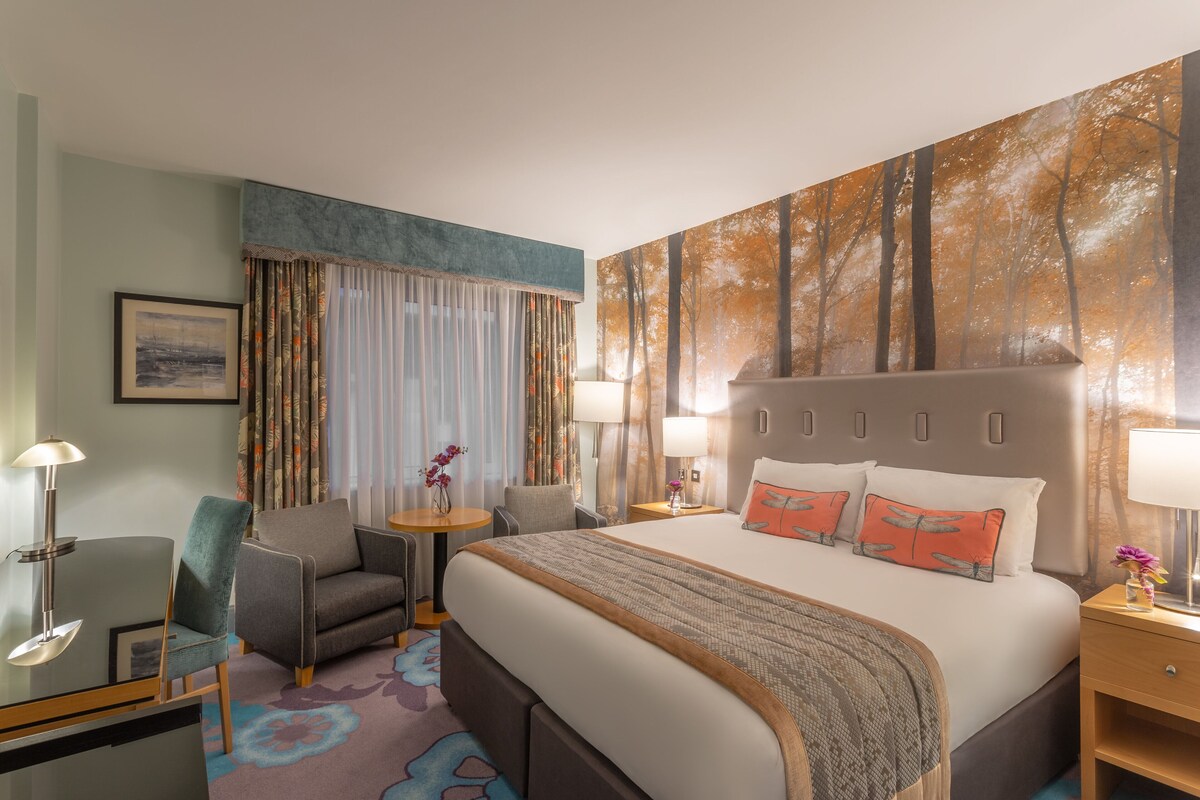 Une chambre d’hôtel avec un grand lit, deux tables de chevet, un bureau avec une chaise, une petite table ronde avec une chaise, et une grande fenêtre avec des rideaux.