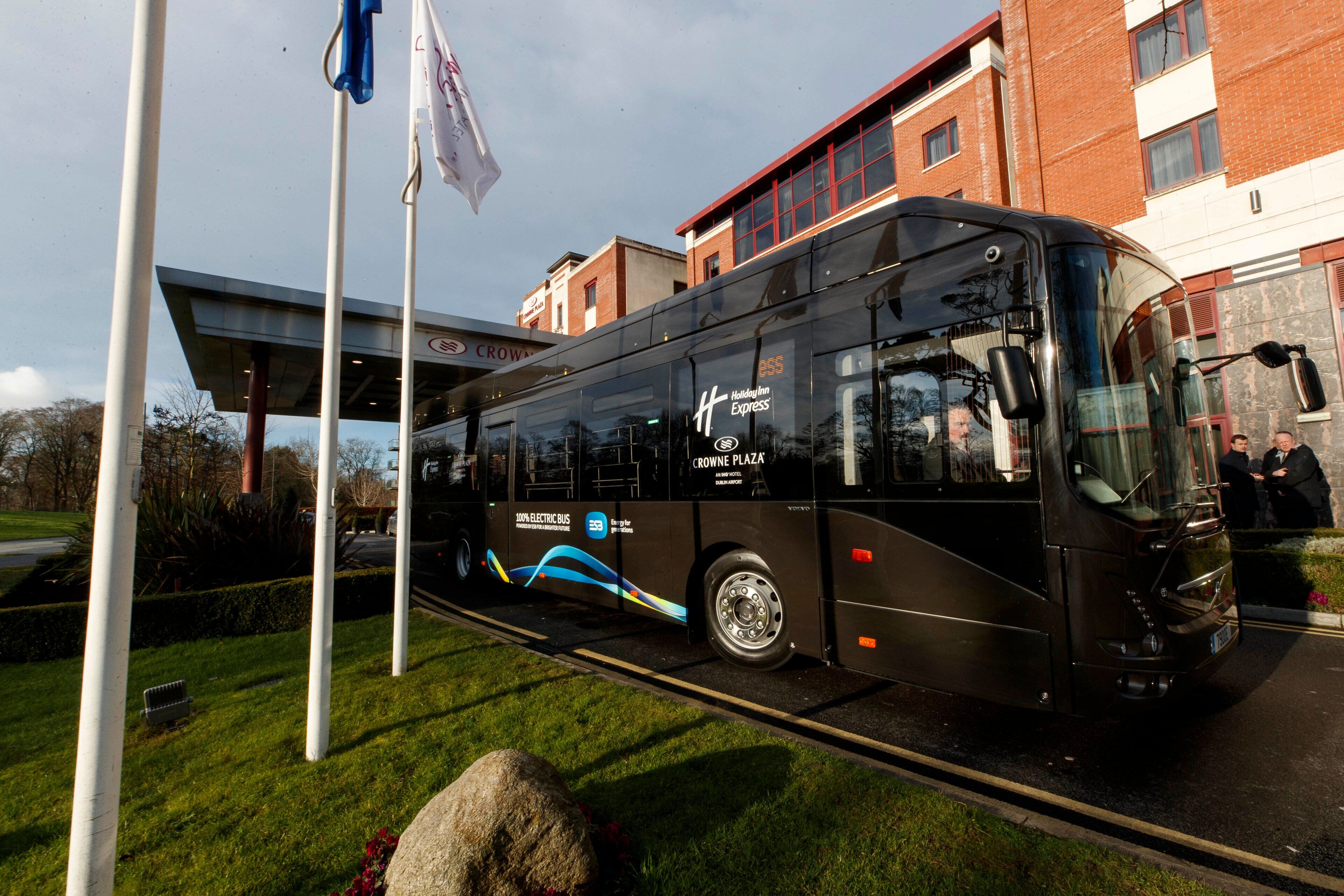 Un autobús eléctrico negro con la marca 'CROWNE PLAZA' y 'Hollander Express' estacionado frente a un edificio de fachada roja y blanca.
