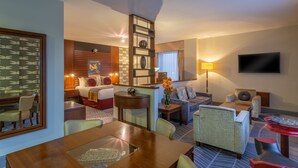 Een hotelkamer met een eetgedeelte, een zithoek met een bank en fauteuils, een aan de muur bevestigde televisie en een groot raam met gordijnen.