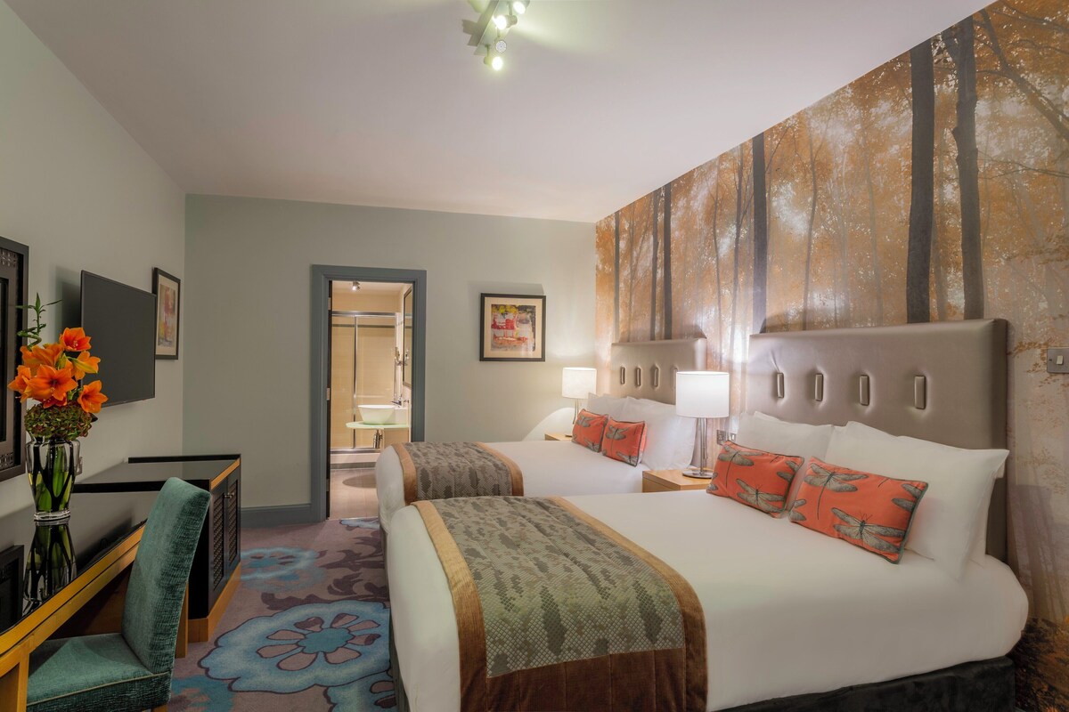 Une chambre d’hôtel avec deux lits, un bureau, une chaise, un vase avec des fleurs et un tableau au mur.