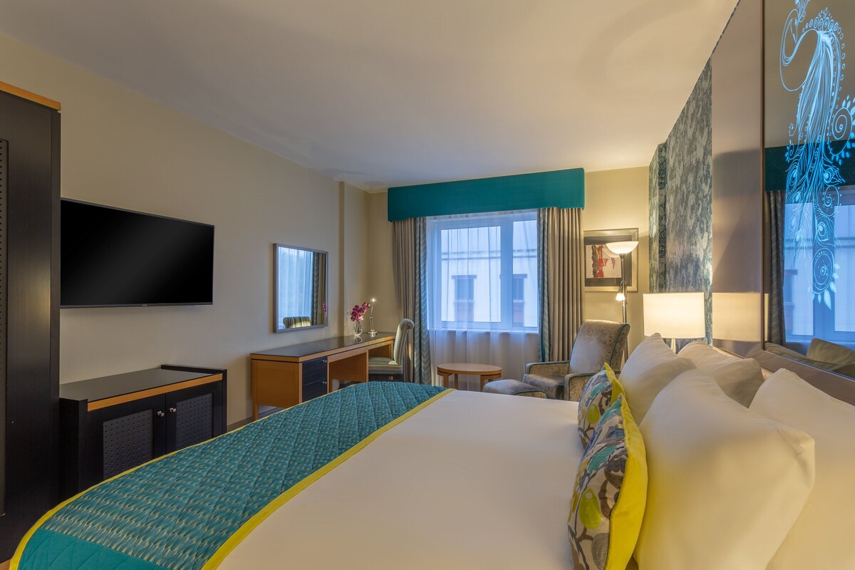 Une chambre d’hôtel avec un grand lit, un bureau avec une chaise, une télévision et une fenêtre avec des rideaux.