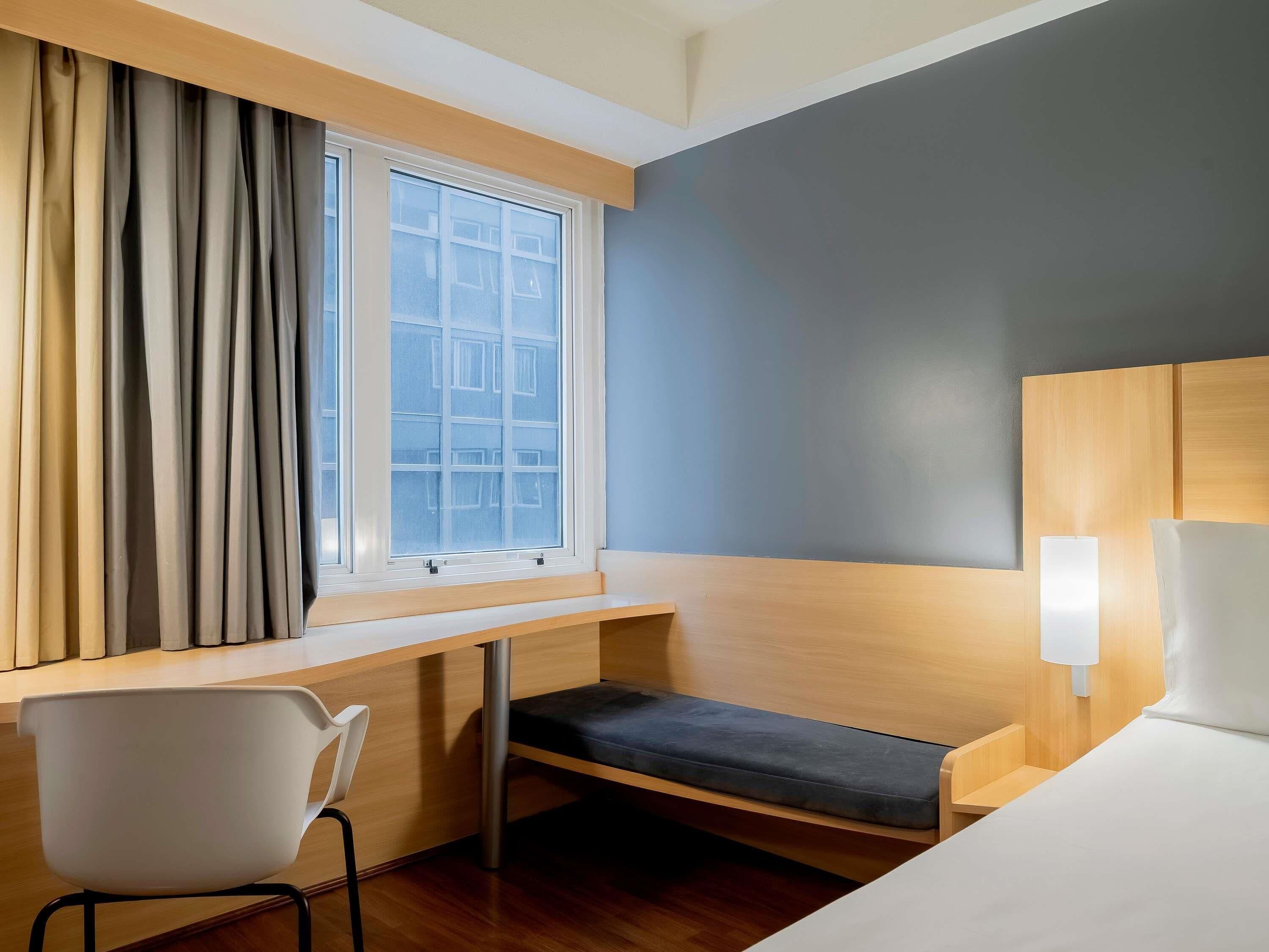 Une chambre d’hôtel équipée d’un bureau, d’une chaise, d’un lit et d’une fenêtre avec des rideaux.