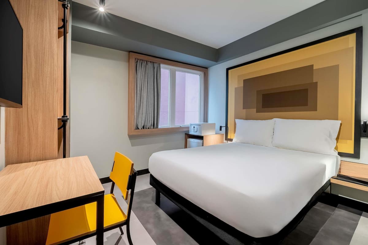 Une chambre d’hôtel comprenant un lit, un bureau, une chaise, une fenêtre avec des rideaux et une table de chevet avec une lampe.