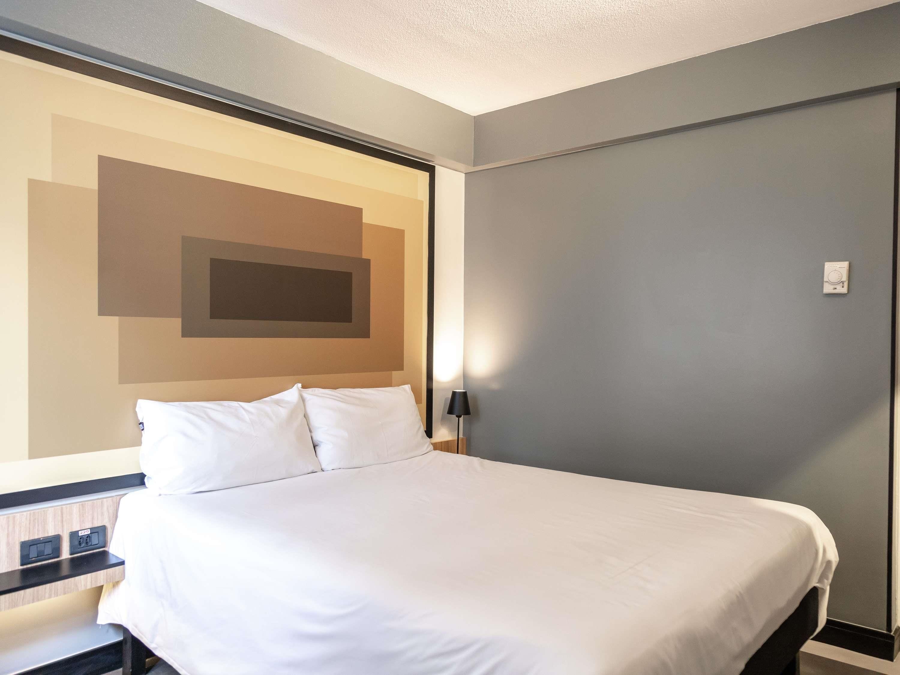 A hotel room with a bed, a nightstand, a wall-mounted lamp, and a thermostat.