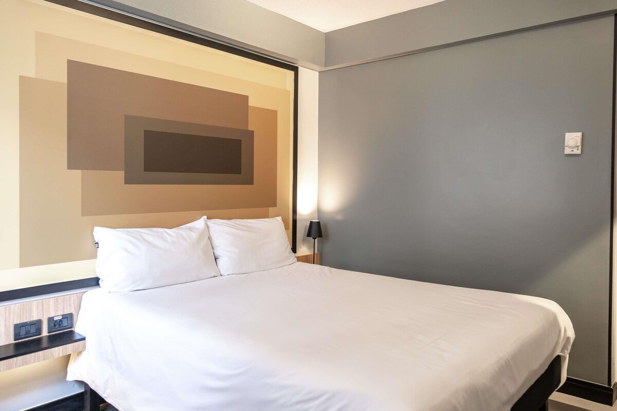 Een hotelkamer met een bed, nachtkastje, wandlamp en een thermostaat.