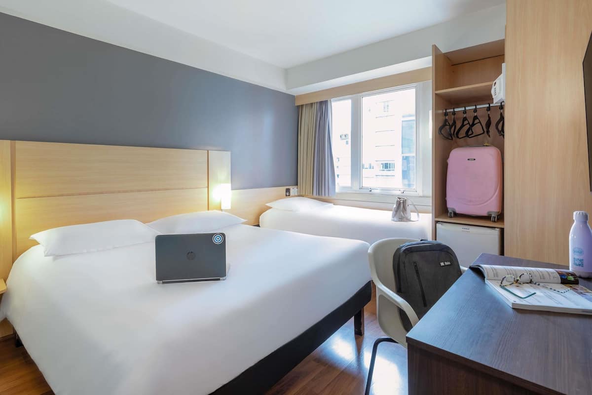 Une chambre d’hôtel avec deux lits, un bureau, une chaise, un ordinateur portable, une valise et une armoire.