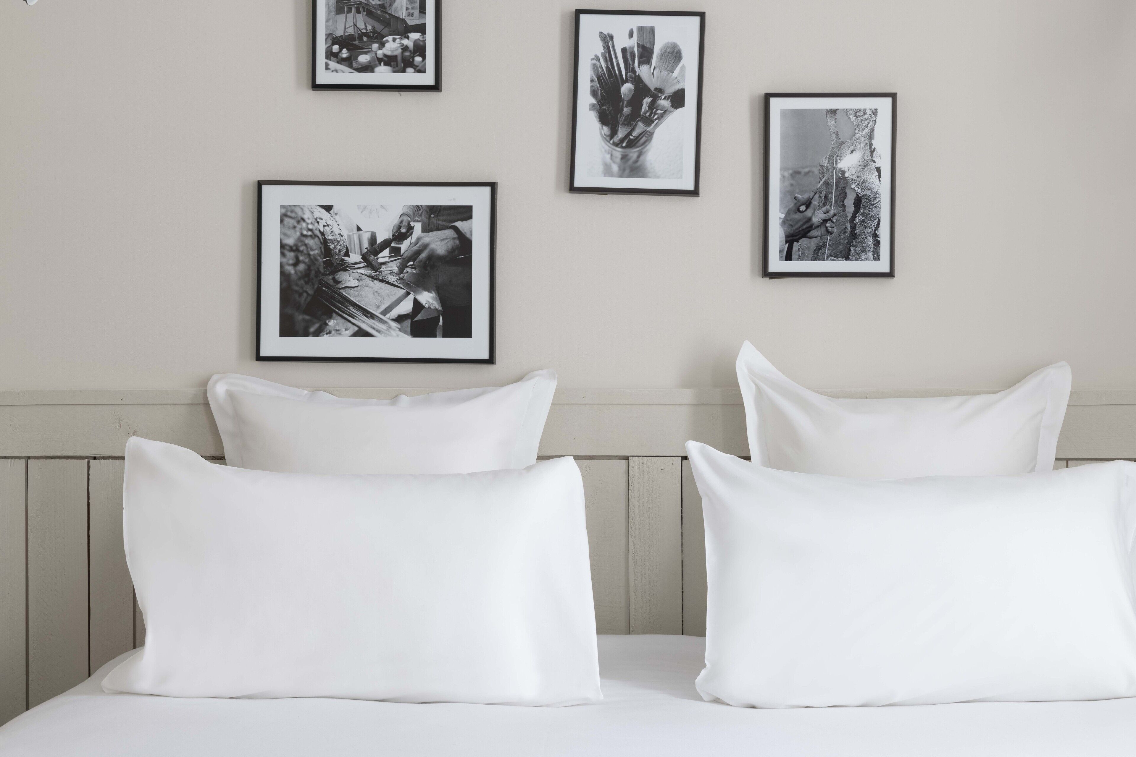 Una cama bien hecha con sábanas y almohadones blancos, fotografías en blanco y negro en la pared y un cabecero de madera.