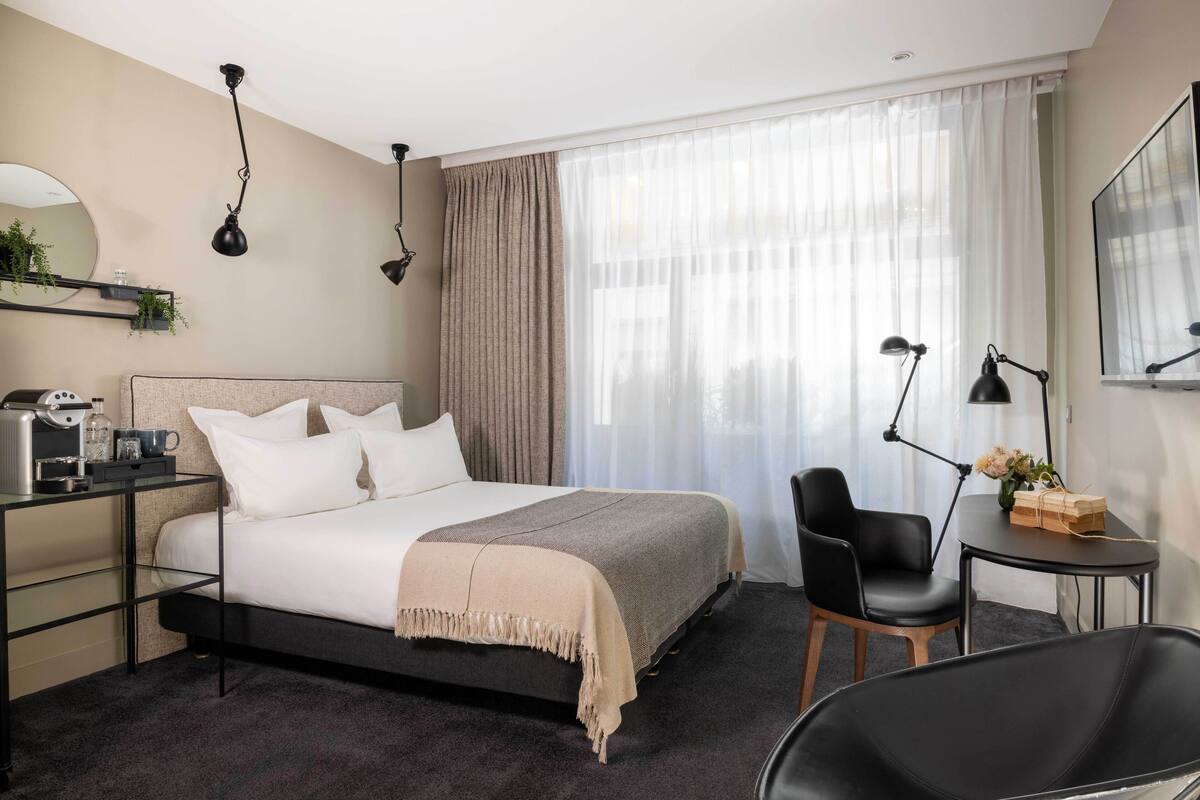 A modern hotel room with a bed, bedside table, chair, and a desk with a lamp.