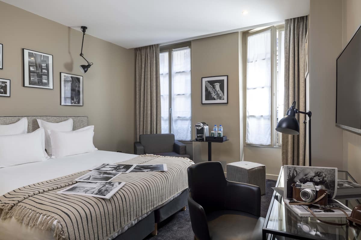 A hotel room with a large bed, a desk with a lamp, a chair, and a table with a camera and a framed picture.