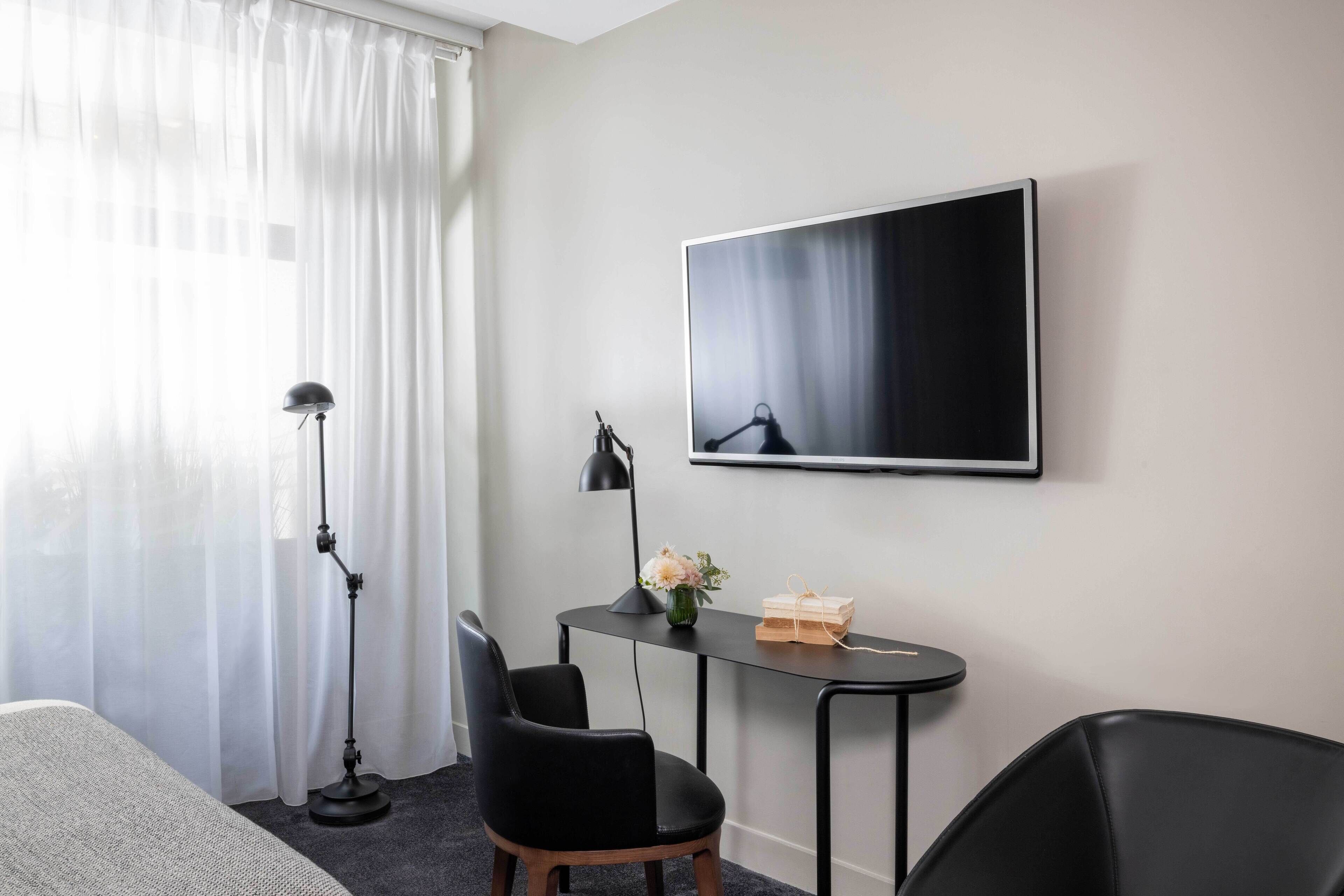 A hotel room with a bed, a desk with a chair, a standing lamp, a flat-screen TV, and a window with sheer curtains.