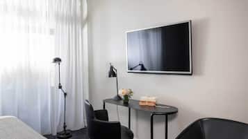 A hotel room with a bed, a desk with a chair, a standing lamp, a flat-screen TV, and a window with sheer curtains.