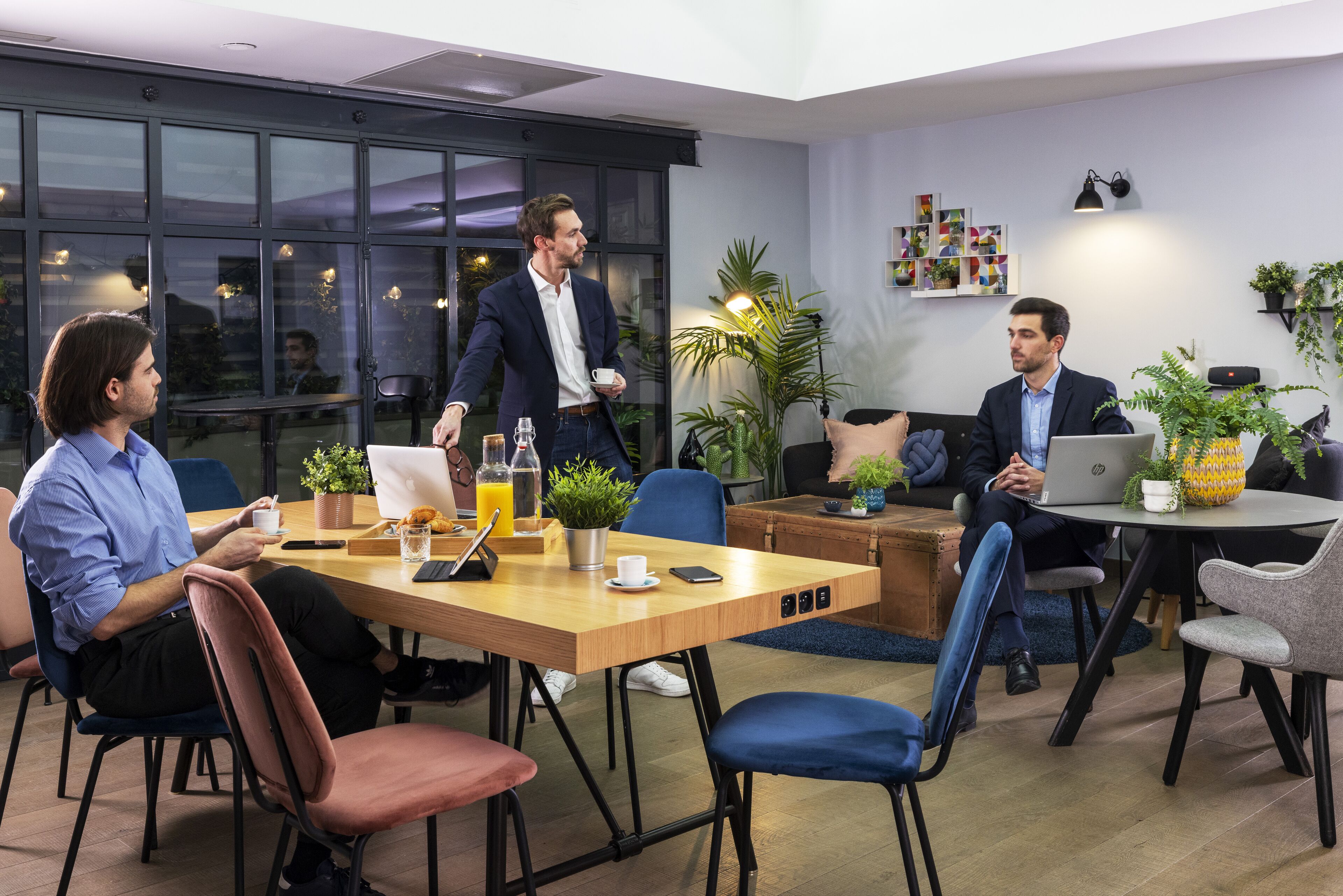 Una sala de reuniones moderna con una mesa de madera, sillas azules y rojas, y un portátil sobre una mesita.