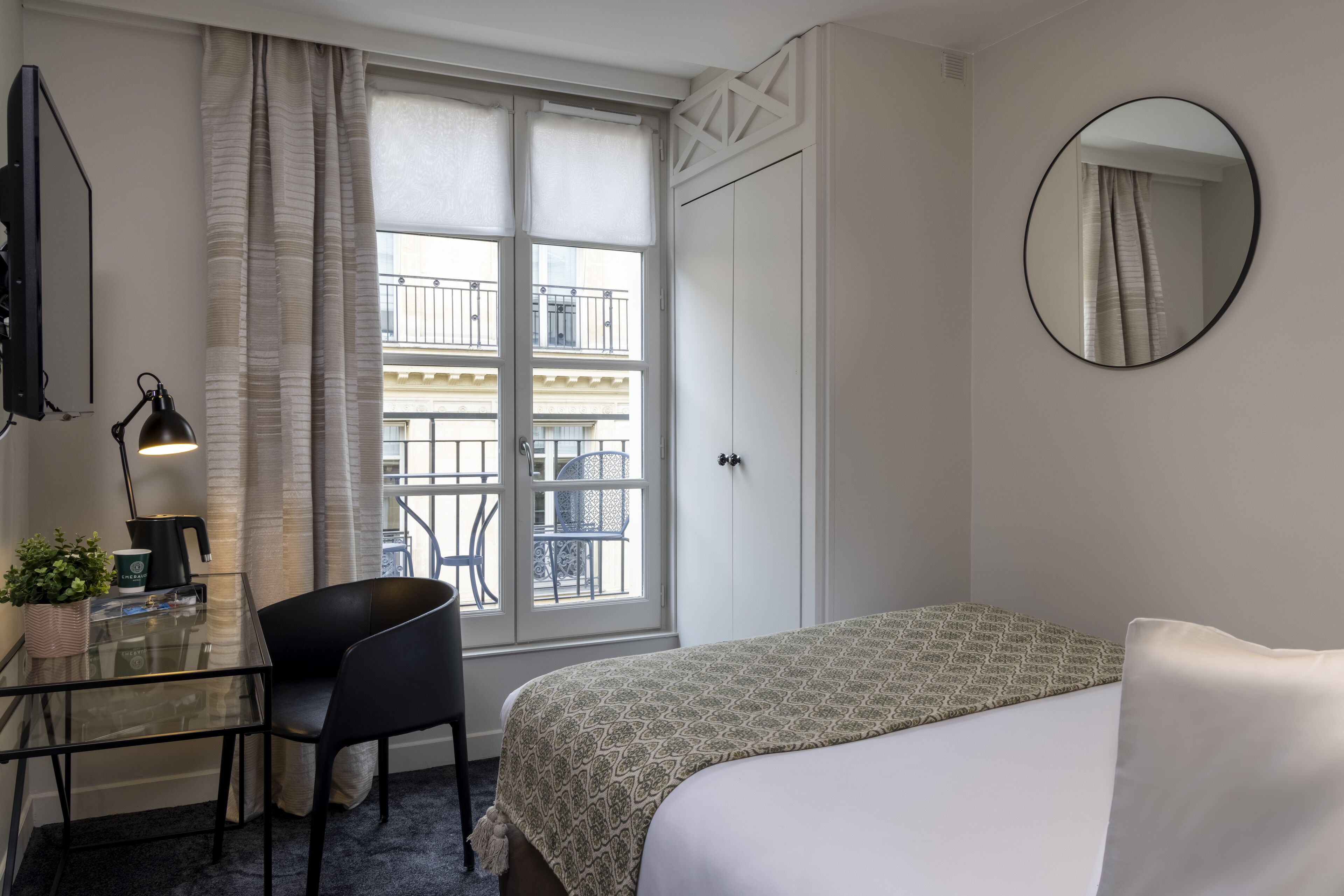 Una habitación de hotel con una cama, un escritorio con una silla, una ventana con cortinas, un espejo redondo y vistas a un balcón con mobiliario exterior.