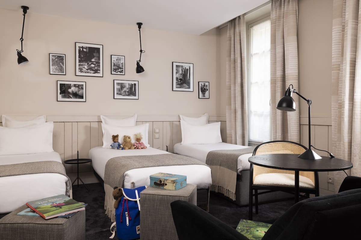 A hotel room with two beds, a small round table, a chair, and a bookshelf.