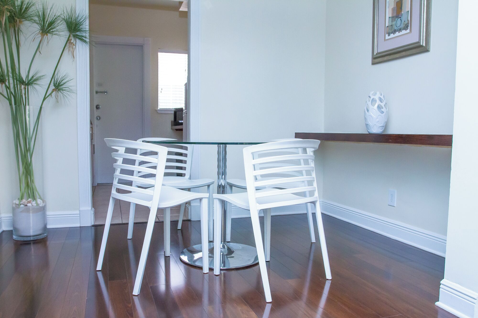 Un coin repas avec une table à plateau en verre et quatre chaises blanches, une plante en pot et un vase blanc posé sur une étagère.