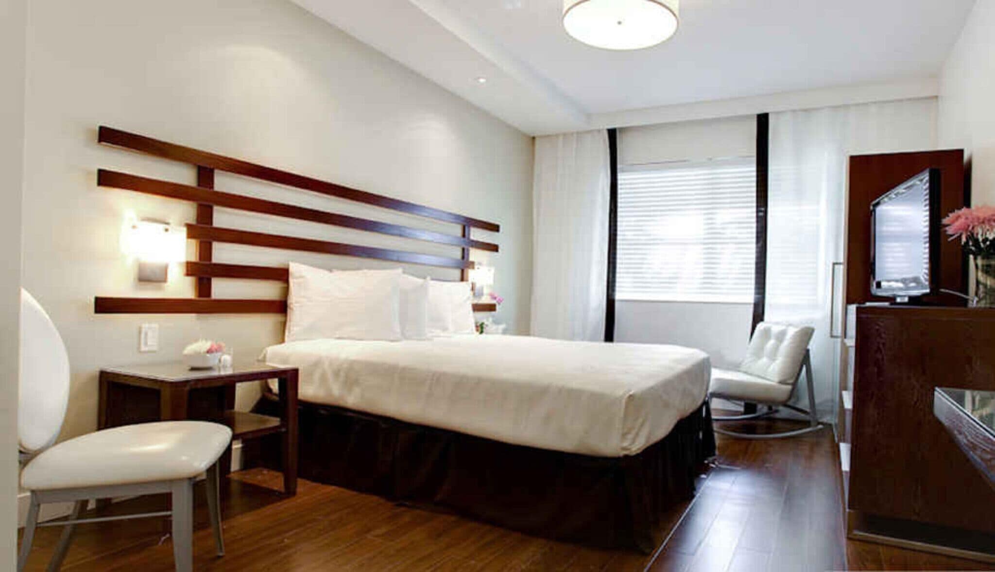 A hotel room with a bed, a desk, a chair, a desk lamp, a mirror, and a window with blinds.