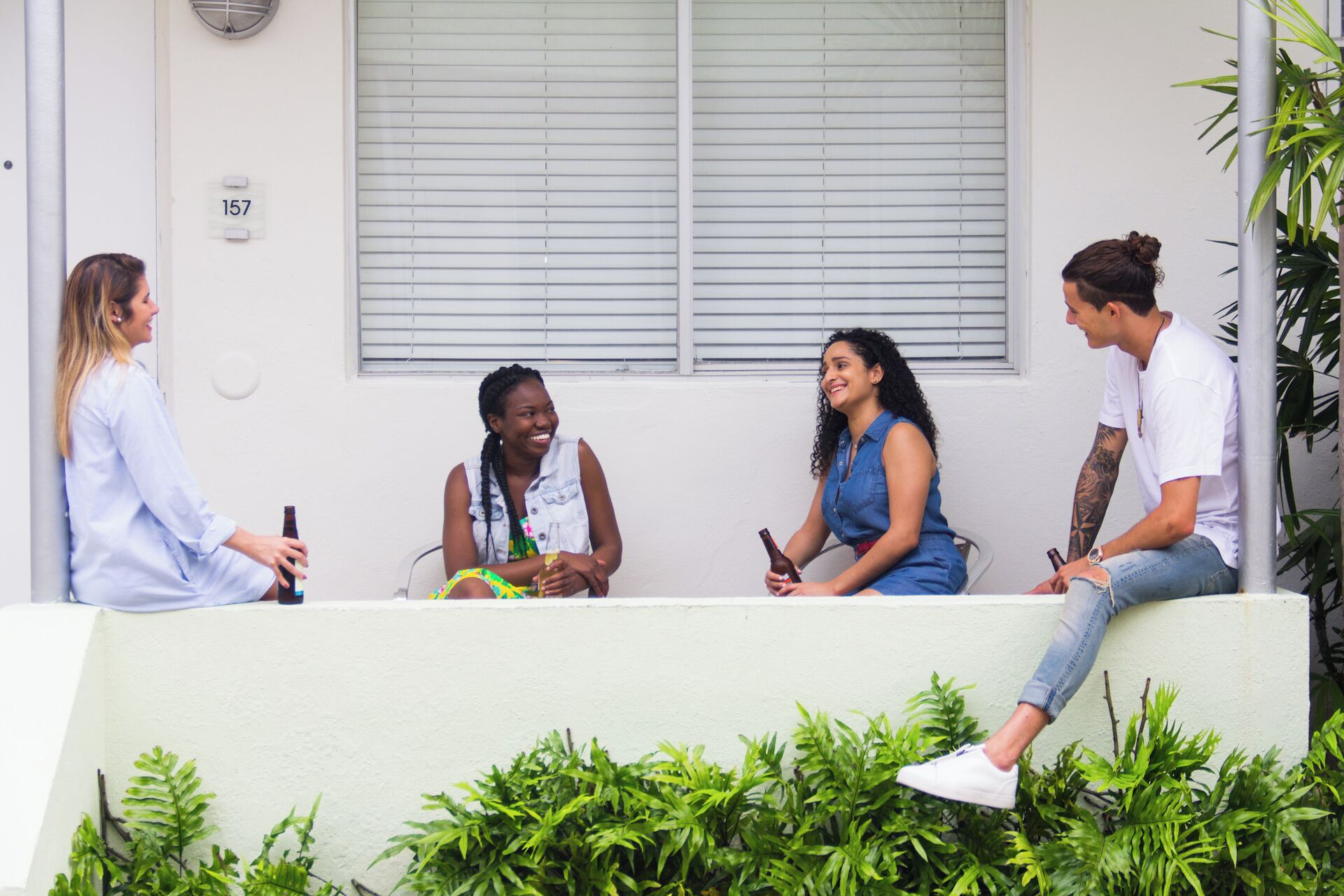 Four people sitting on a white outdoor bench in front of a building with a window and a door marked with the number 157.